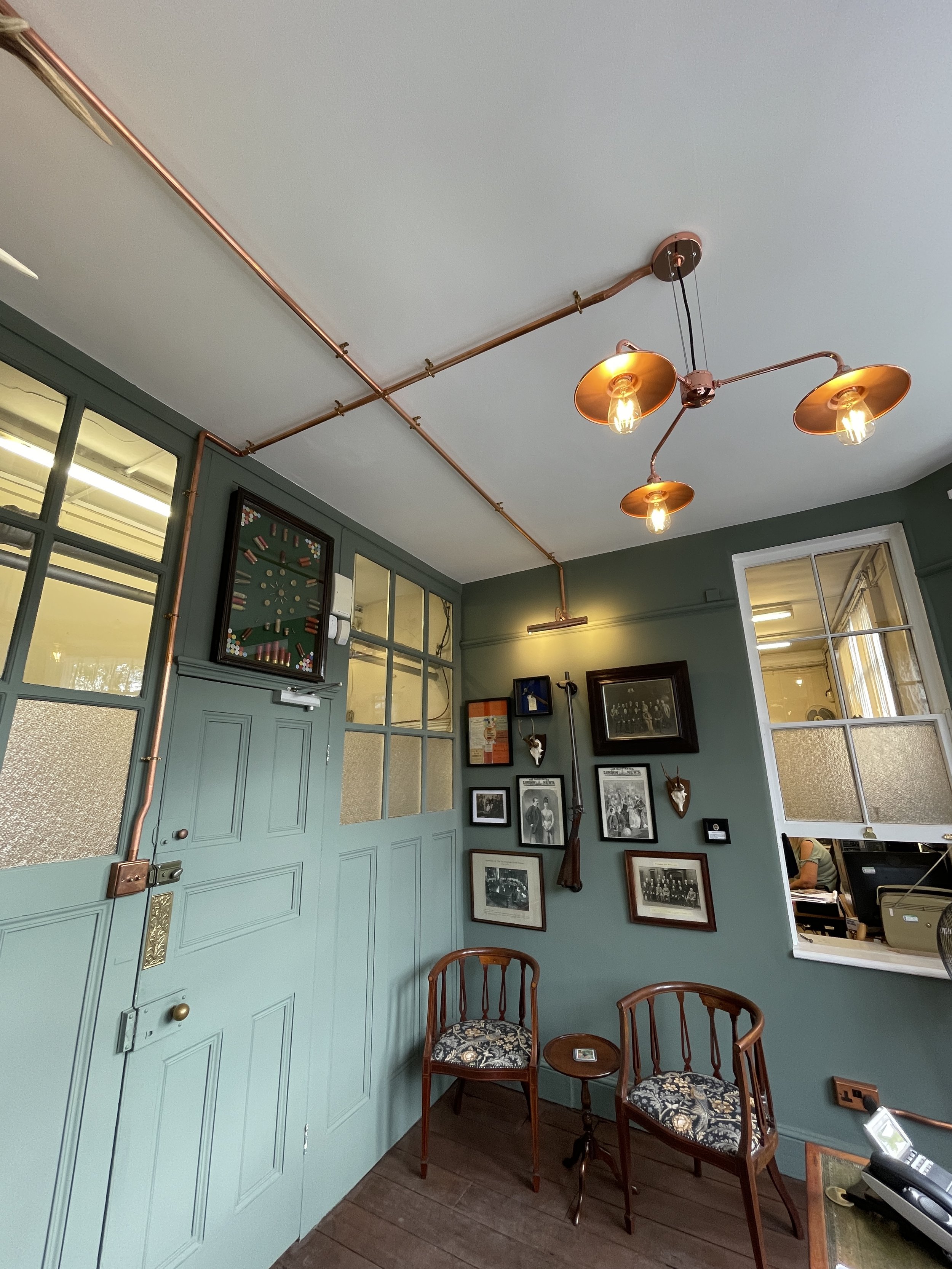 Vintage-style cafe interior with green walls, framed photos, and eclectic decor, featuring two wooden chairs with floral cushions and a small wooden table between them.