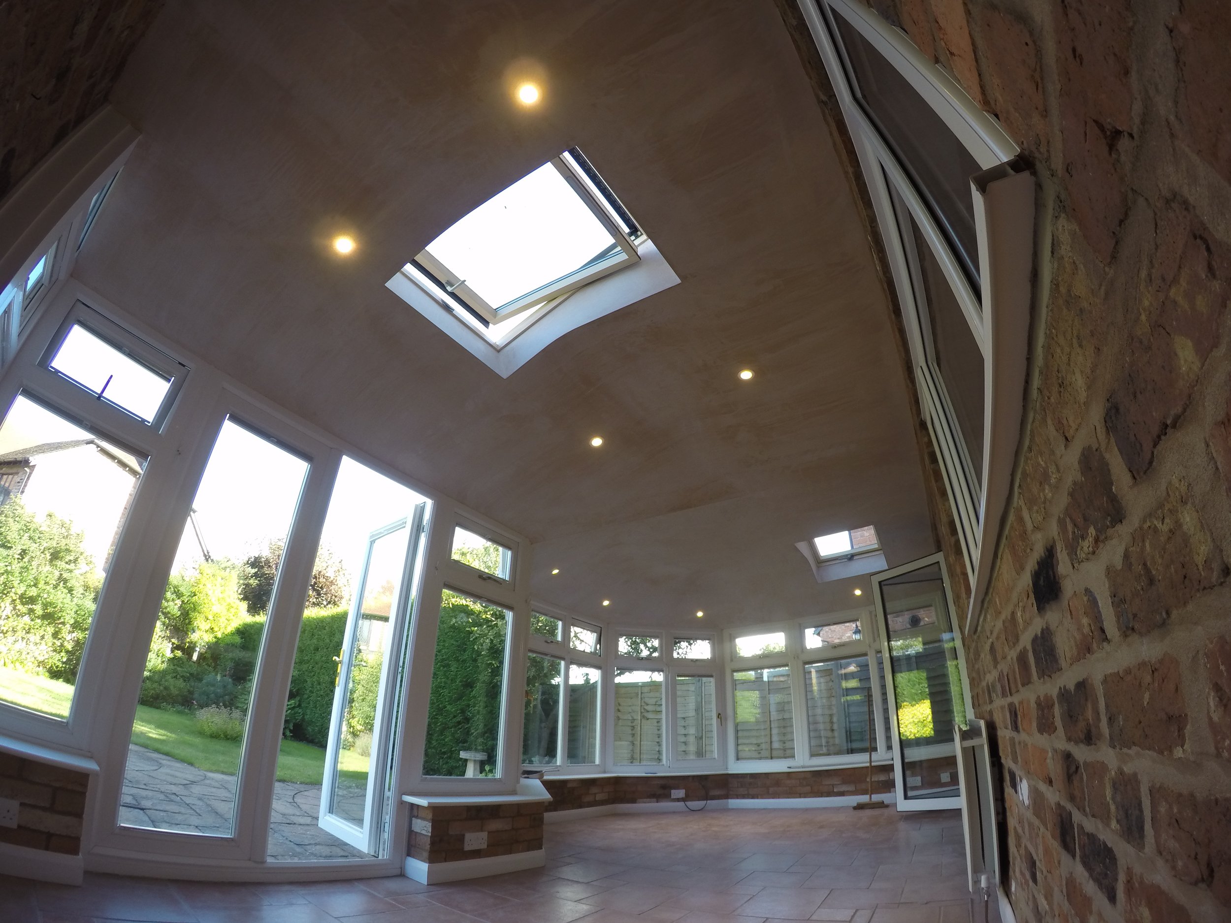 Bright sunlit enclosed patio with large windows, a skylight, and brick walls. Outside garden and greenery visible through the windows.