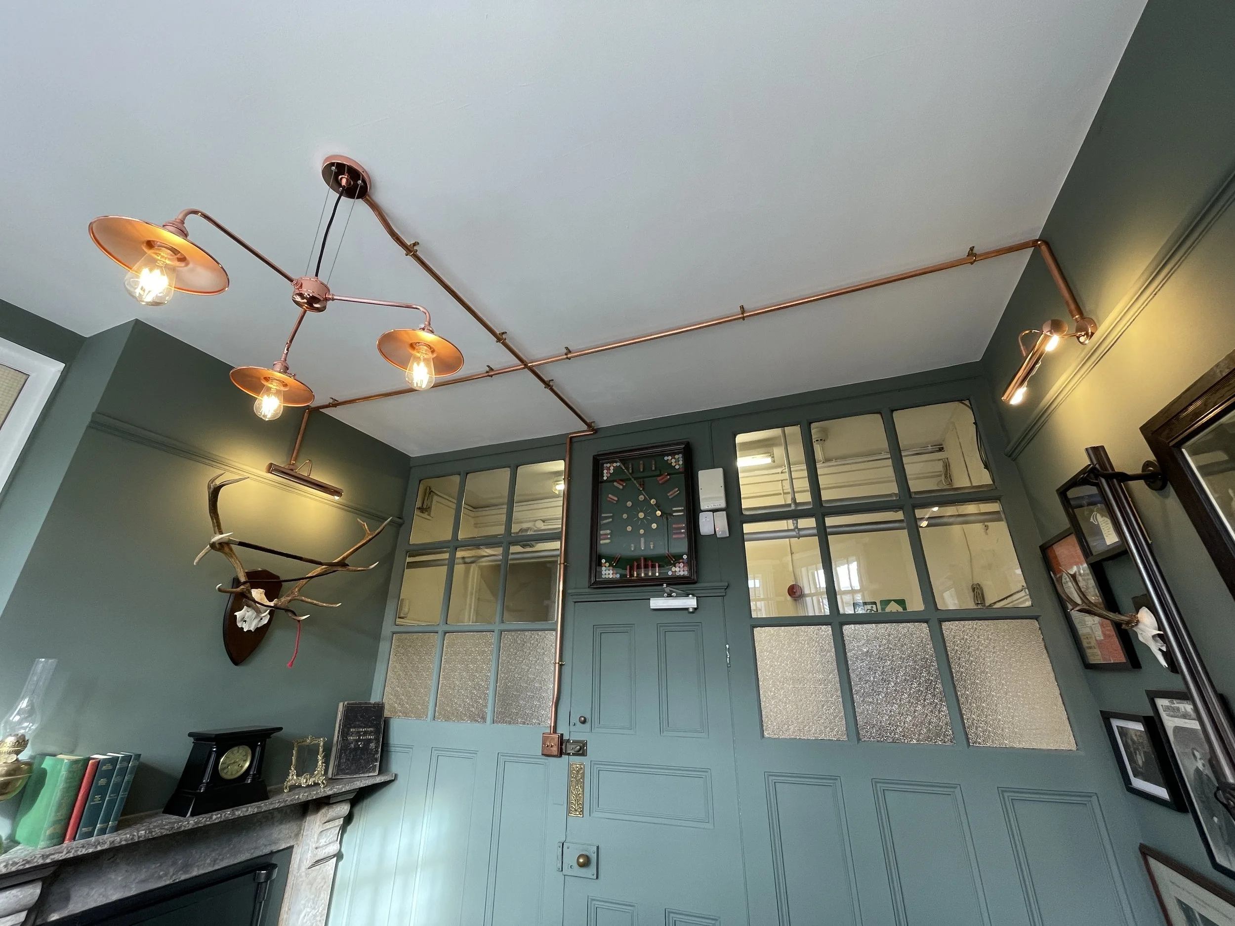 Interior of a room with green walls, featuring vintage-style ceiling lights with exposed bulbs and a copper pipe installation, a mounted antler display, and various framed pictures and clocks.