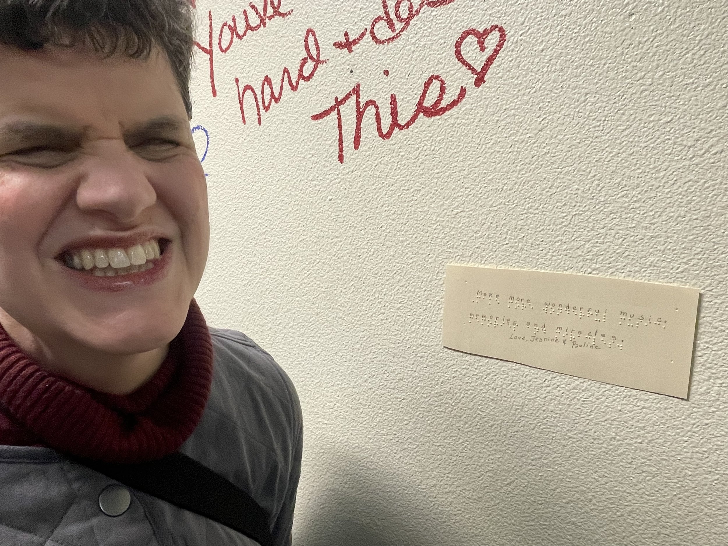 Jeanine Mooers smiling next to a note she wrote in braille saying, "Make more wonderful music, memories, and miricales."