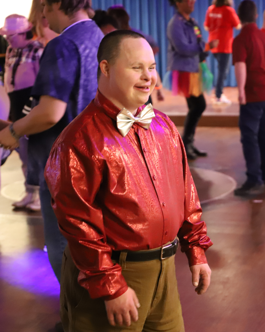 A person with a disability, possibly with Down syndrome, smiling at a dance or party, wearing a shiny red shirt, khaki pants, and a gold bow tie, surrounded by other people in colorful attire.