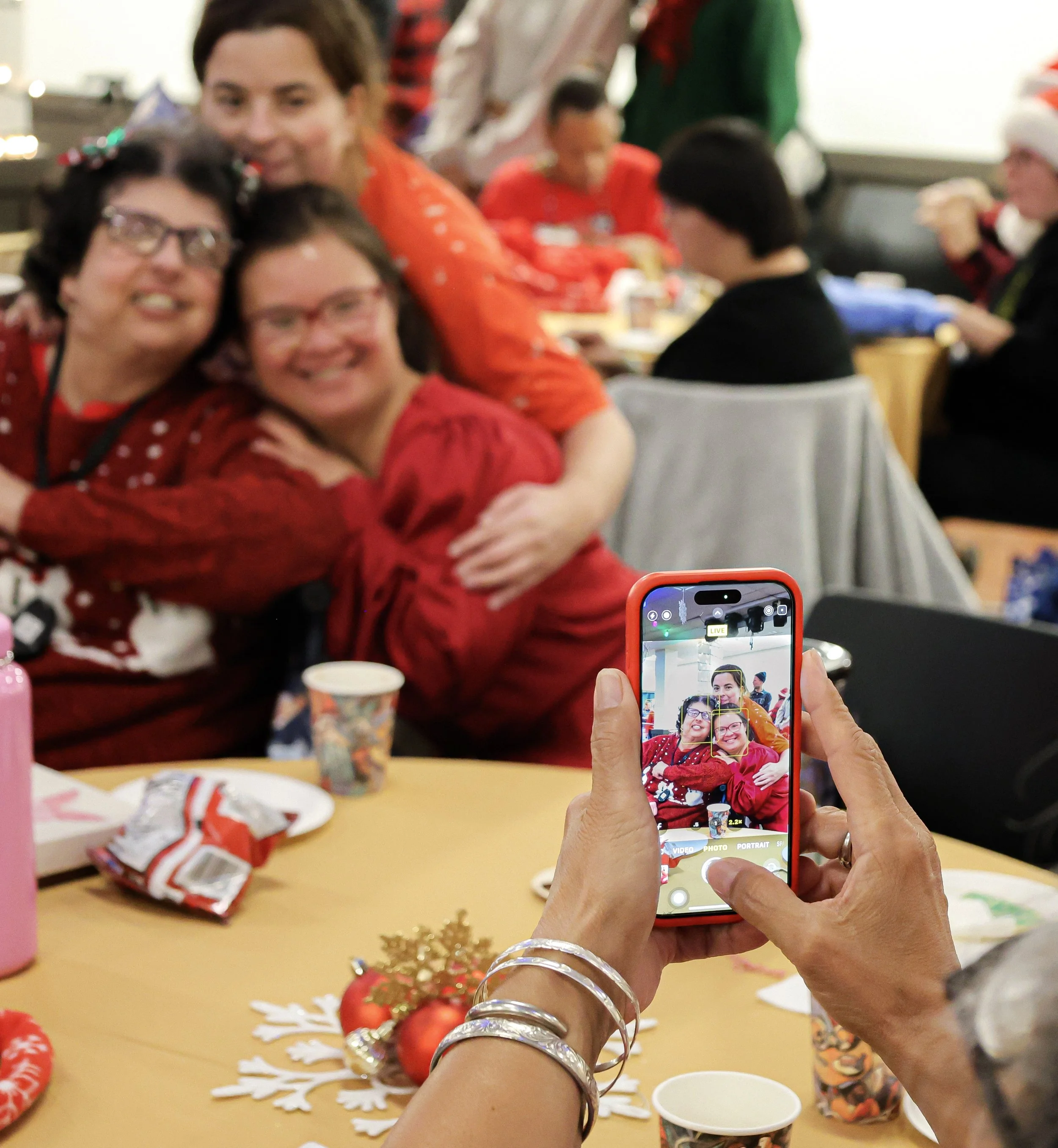 Group of three women hug and smile while someone takes their photo with an iphone