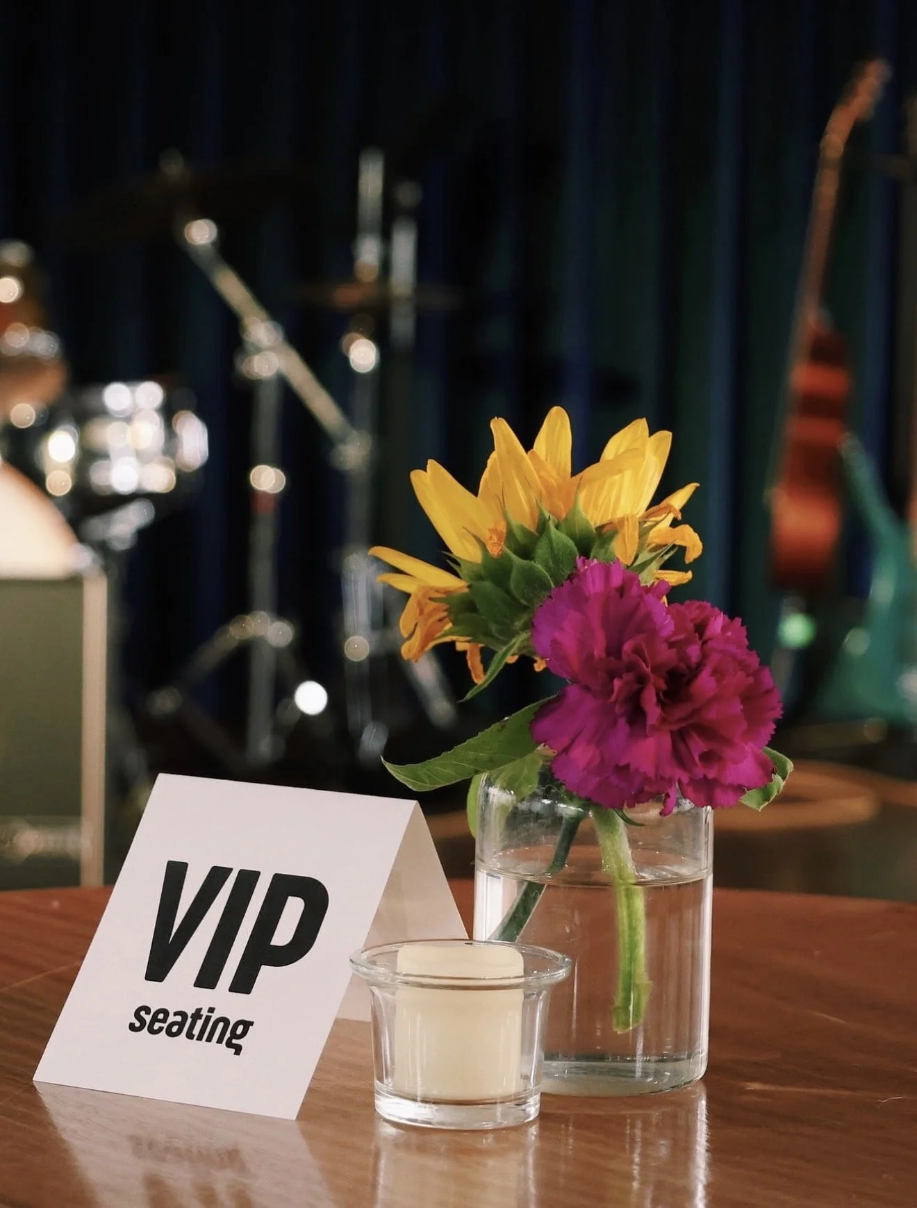 A table with a 'VIP seating' sign, a small candle holder, and a glass vase with yellow and pink flowers in water, with musical instruments blurred in the background.