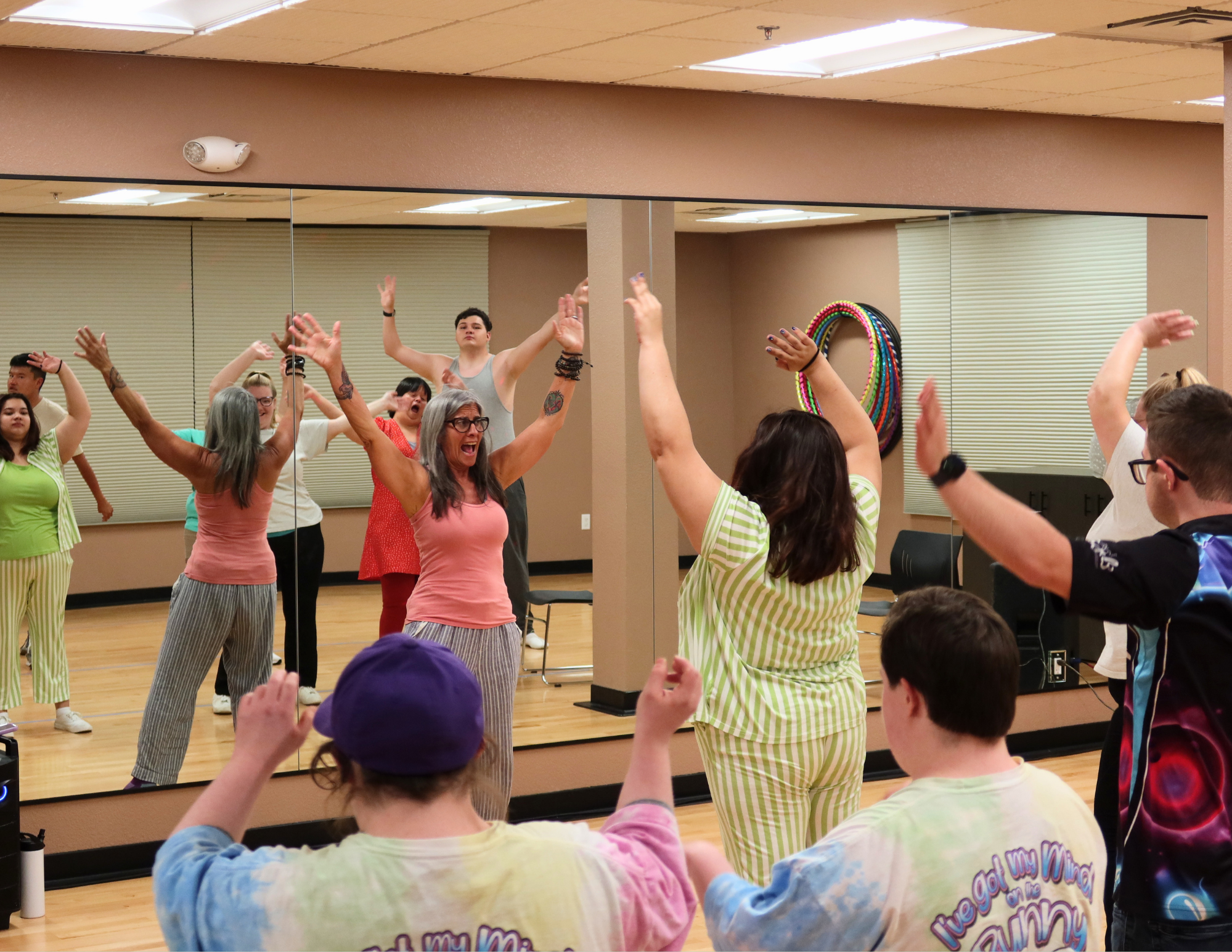 Volunteer leading NMTS dance class in warm ups, volunteer and participants smile with arms raised in the air.