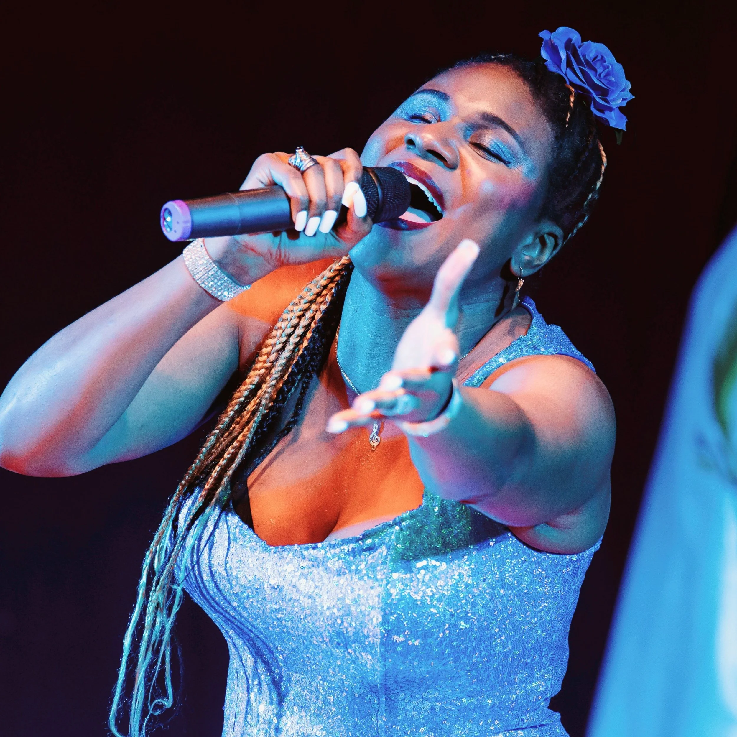 Artist Lachi sings passionately into a microphone on stage, wearing a sparkly blue dress and a blue flower in her hair, reaching one hand toward the audience under colorful stage lighting.