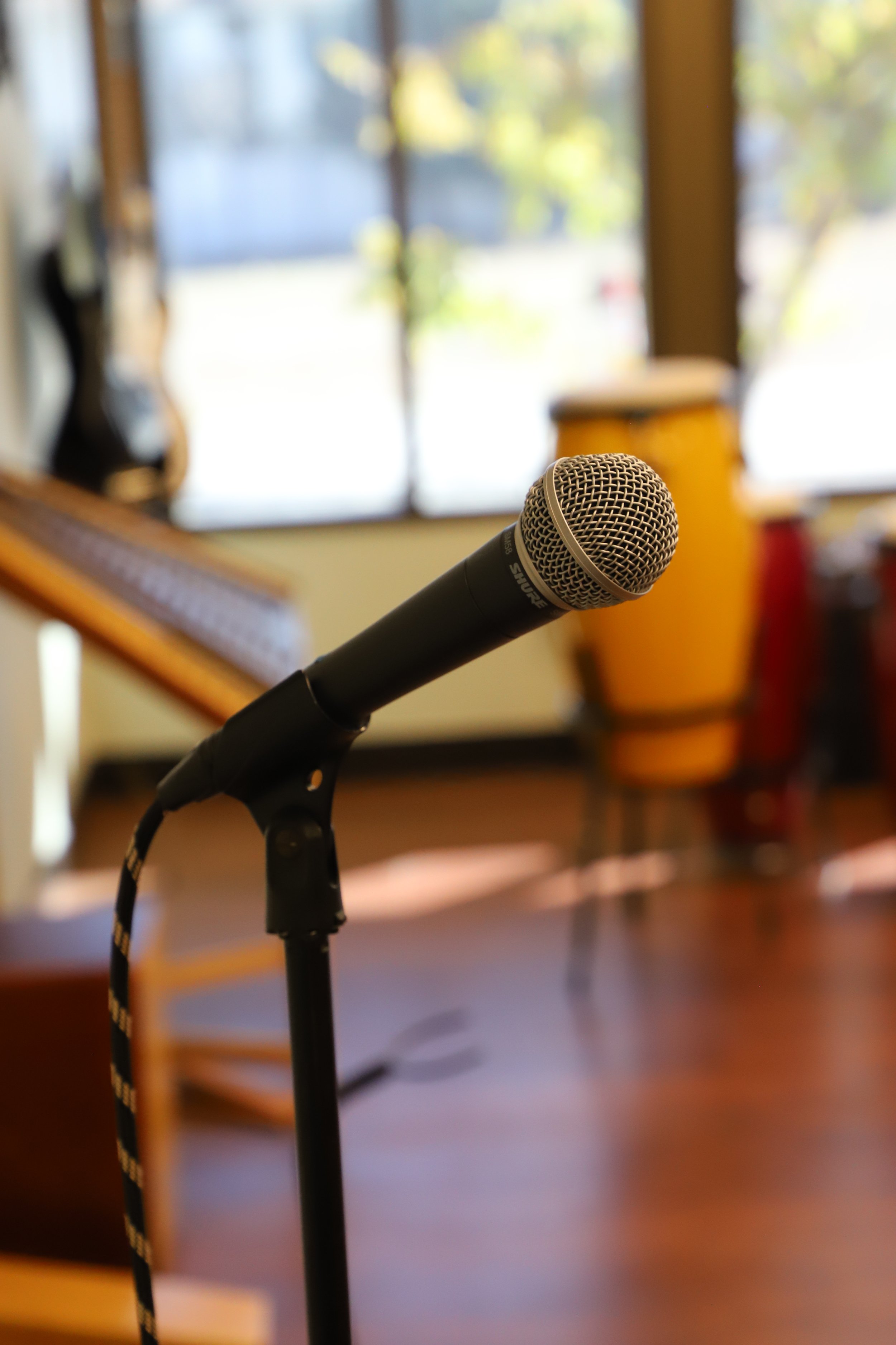 Close-up of a Shure microphone on a stand, set up indoors with a blurred background of a window and musical instruments.