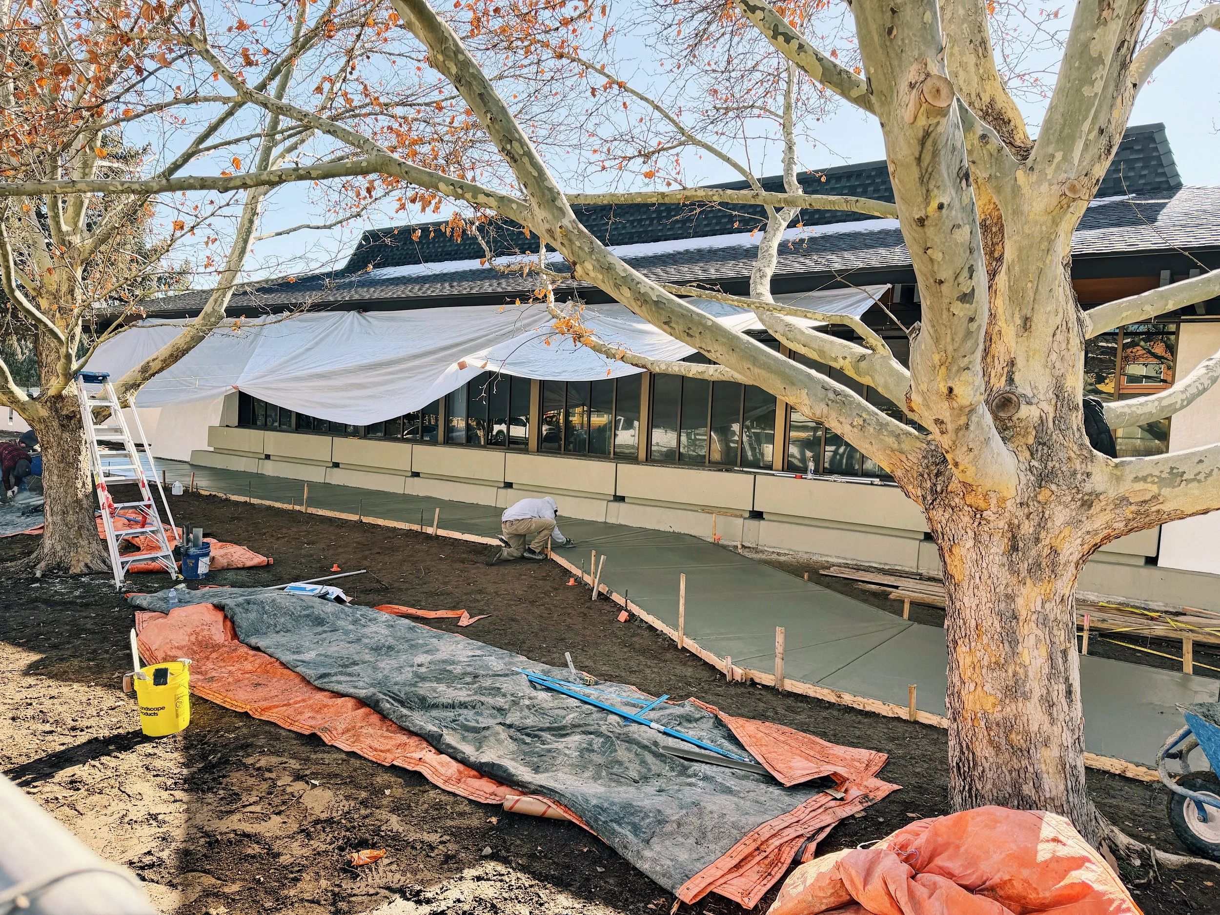 Photo of the front of the NMTS building during construction. Fresh concrete has been poured in front of large front-facing windows, and a worker is on the ground smoothing the surface. Large orange tarps lie nearby.