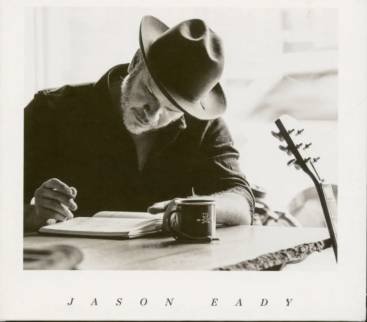 Black-and-white photo of singer-songwriter Jason Eady wearing a hat, seated at a table writing in a notebook, with a guitar resting nearby.