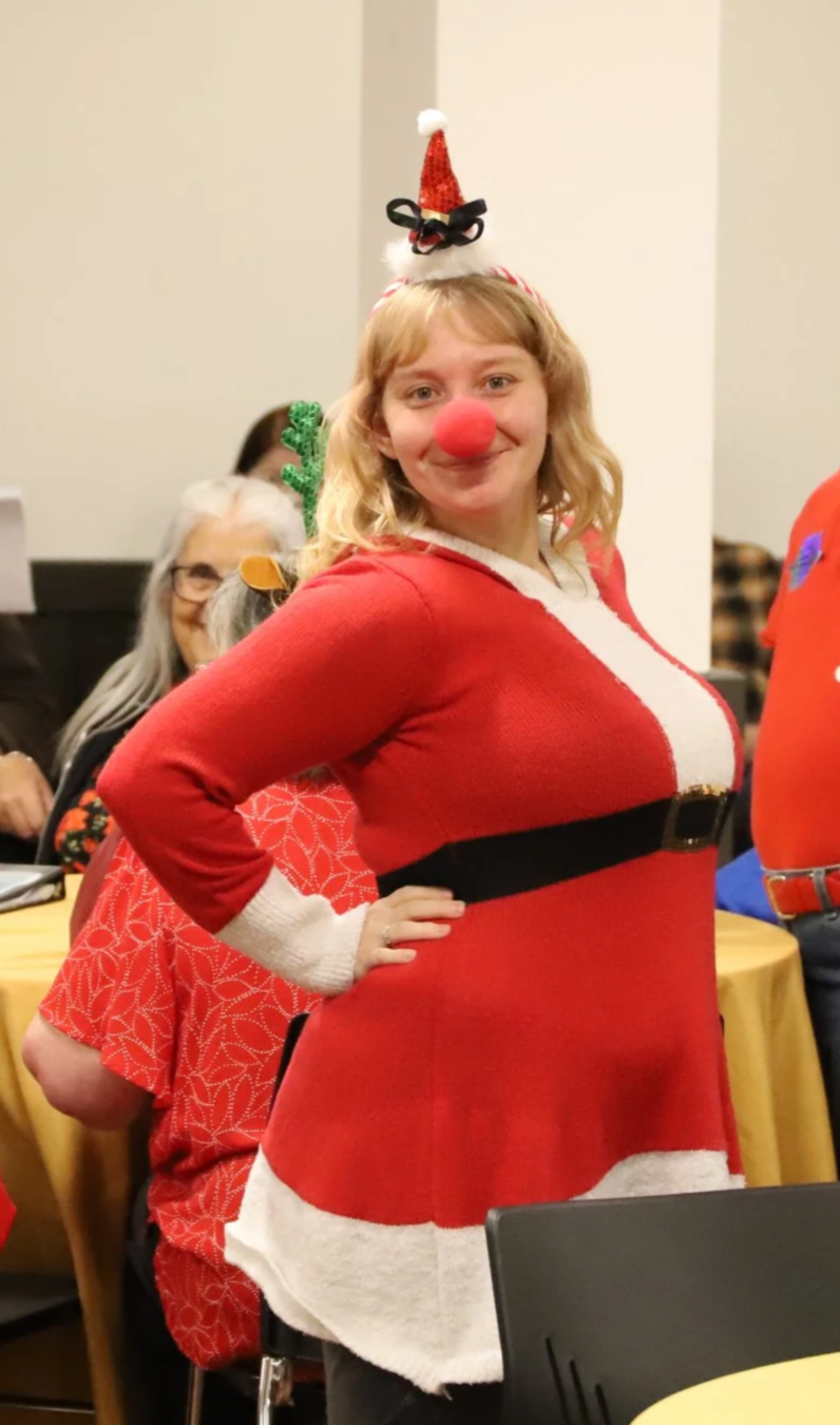 Girl dressed as Mrs. Claus with a Santa hat headband and red Rudolph nose smiling at the camera at the NMTS holiday party.