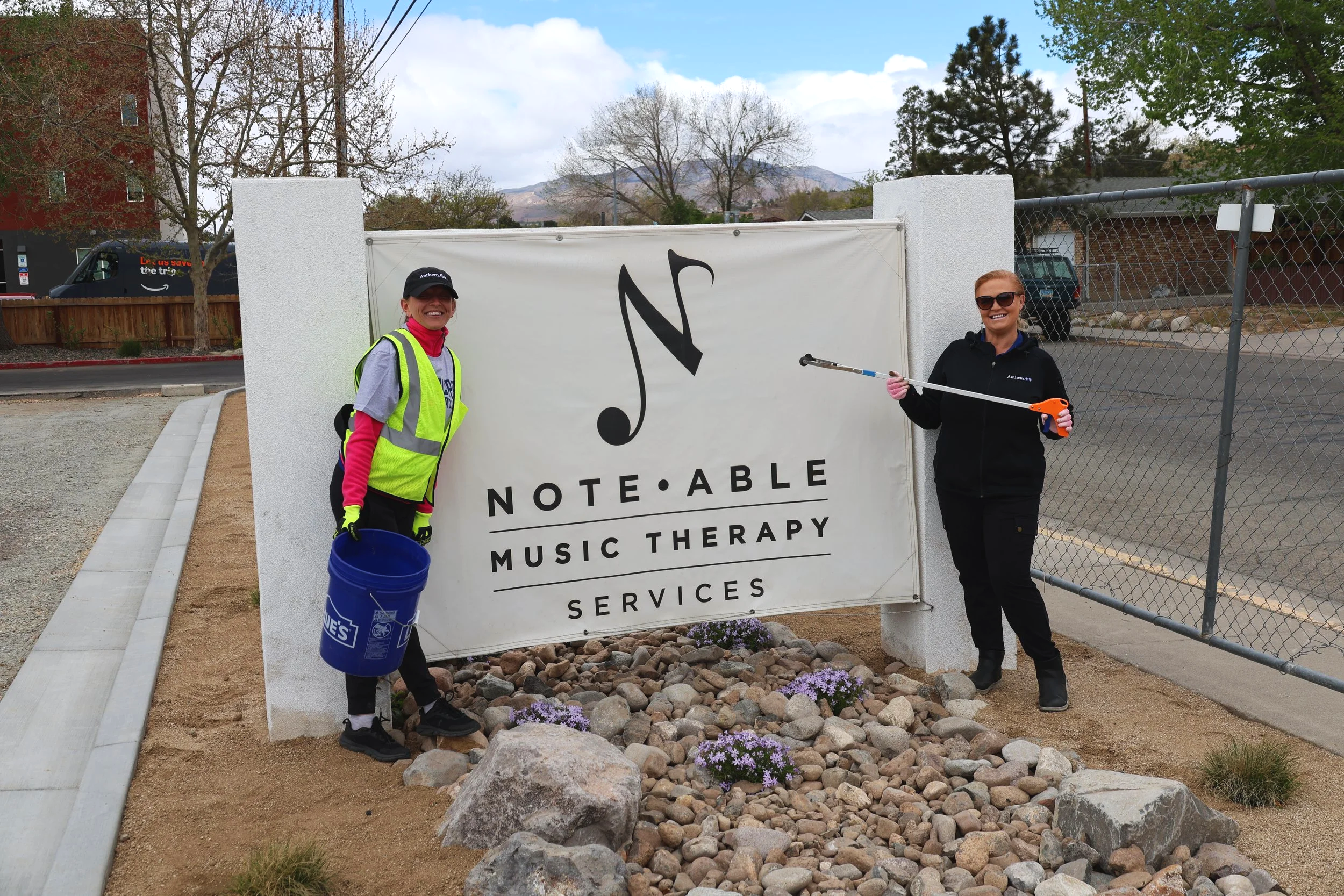 Two volunteers standing in front of the NMTS sign, one holding a bucket and one holding a grabber tool