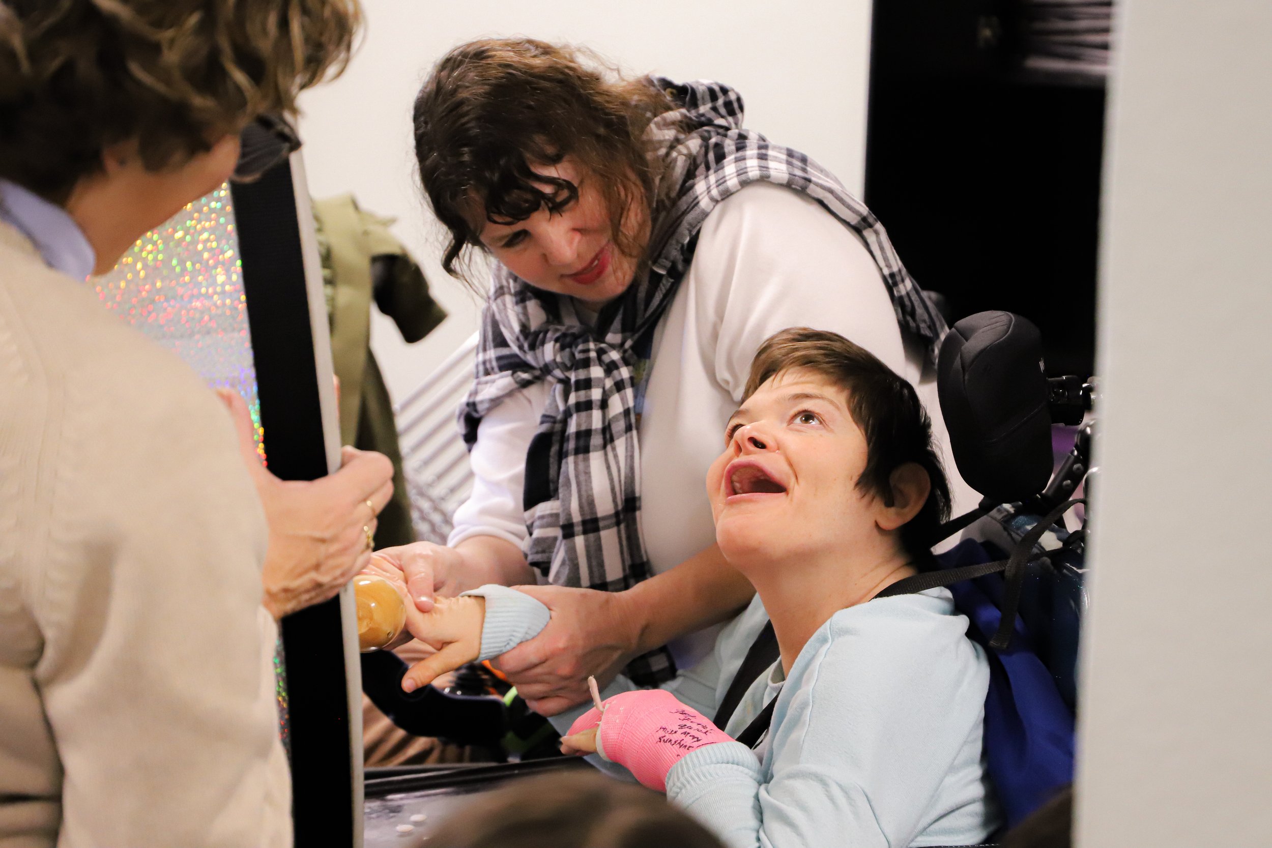 A woman leaning over a young girl in a wheelchair, holding her hand, while another person takes a photo.