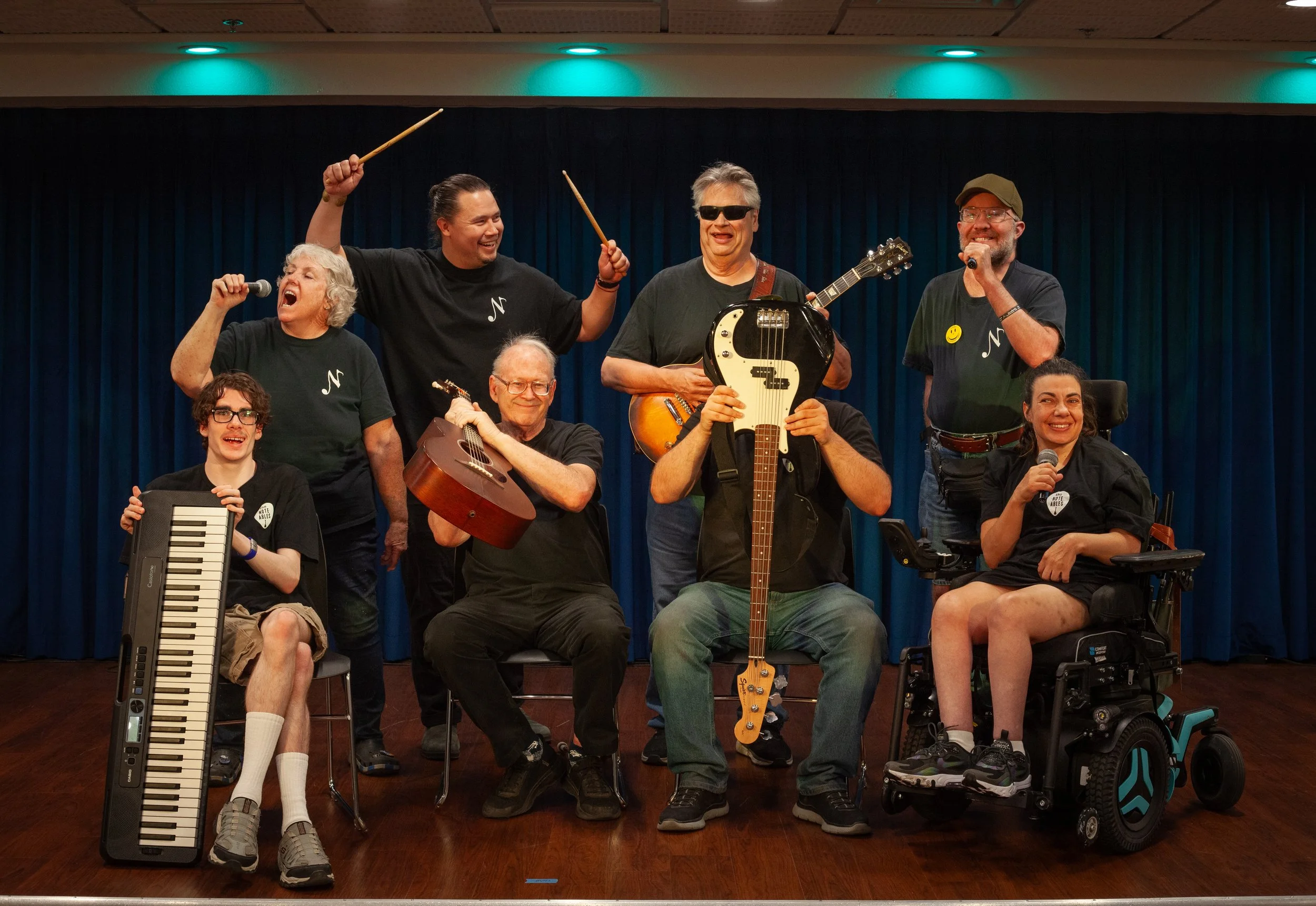 The Note-Ables band members smiling and posing together on stage with musical instruments.