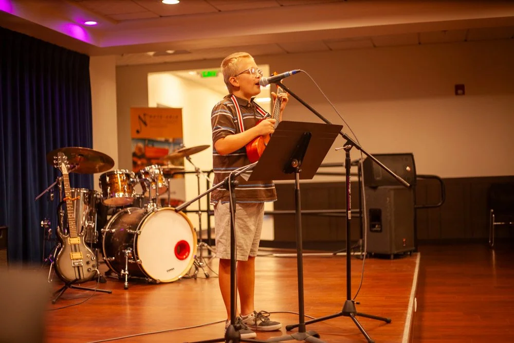 Photo from 2024 NMTS Spring Recital with young participant on stage singing into microphone with Ukulele in hands.