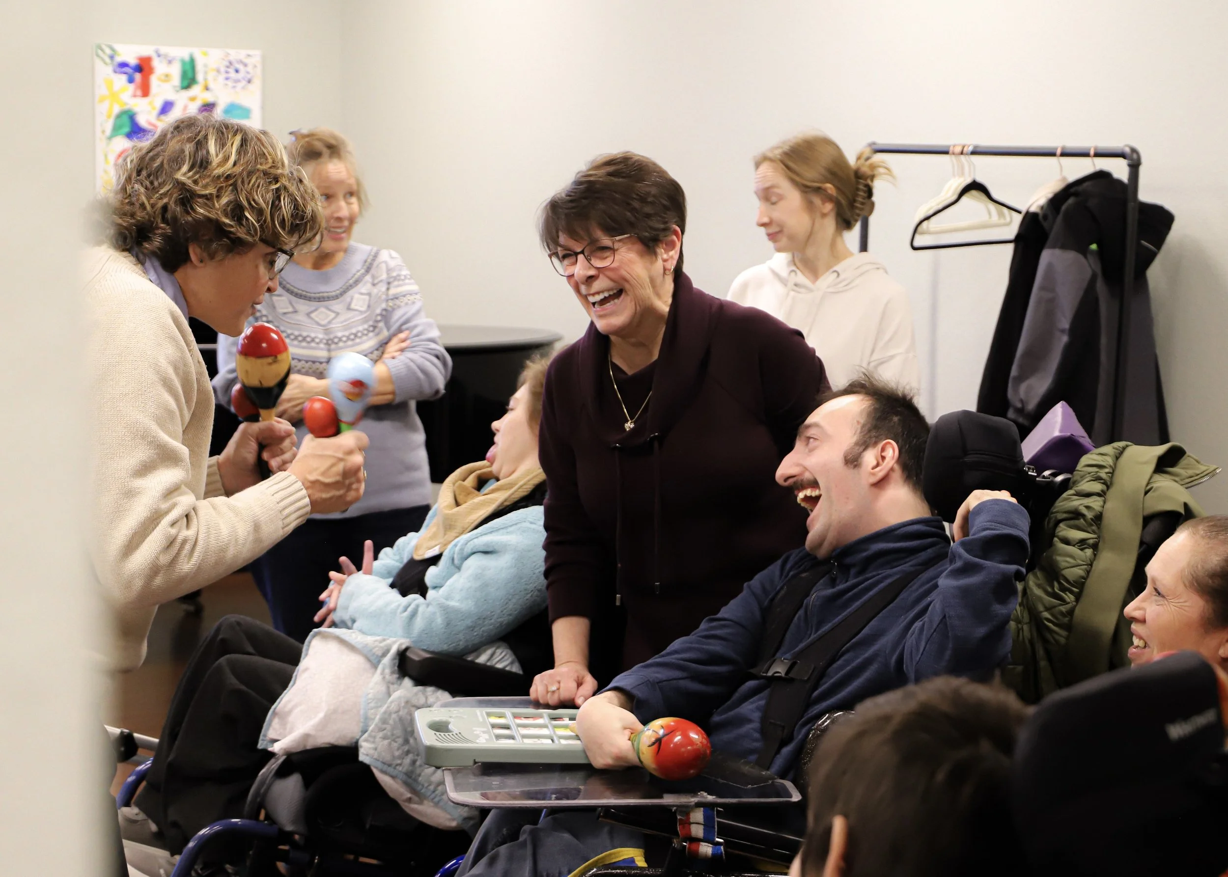 A group of people, some in wheelchairs, laughing and playing with colorful maracas in a room with a light-colored wall and artwork in the background.