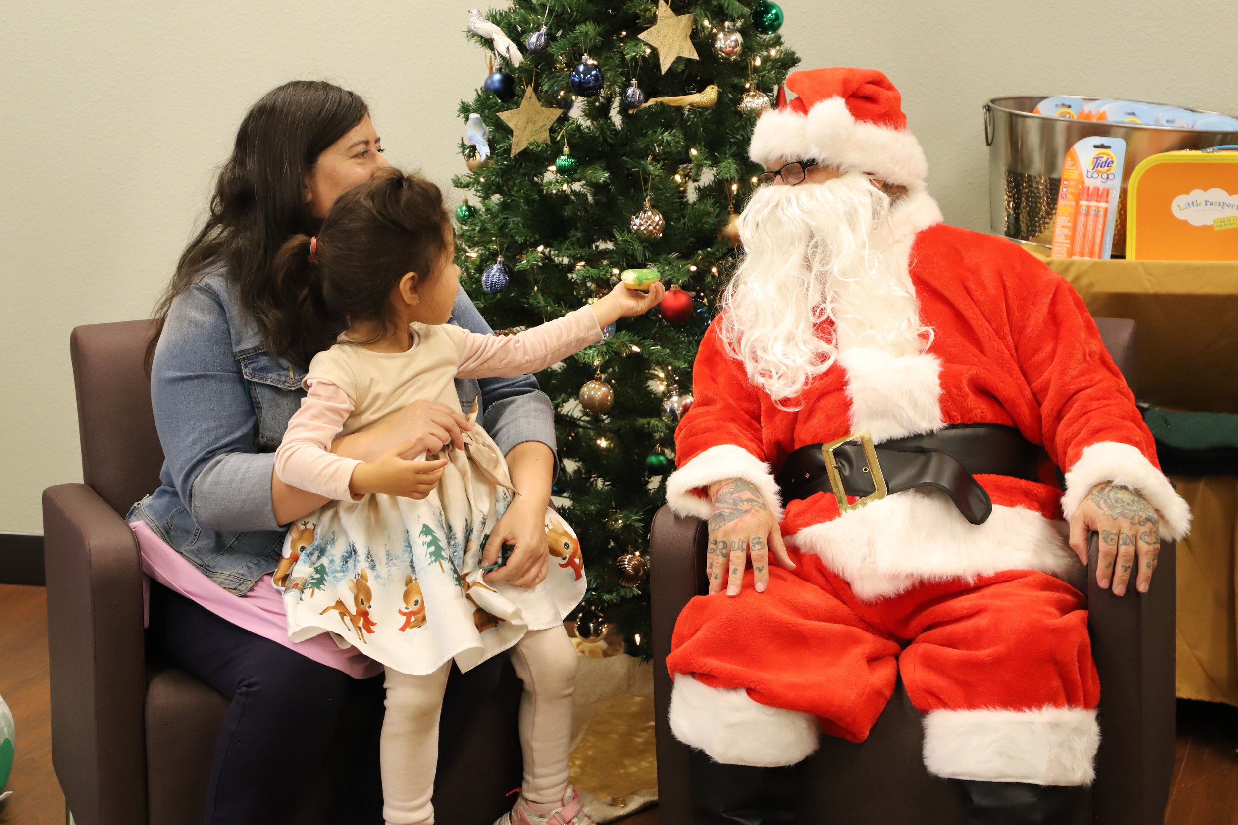 A mom and young daughter sitting with Santa in front of a Christmas tree; the child reaches towards him to show him something she is holding in her hand.
