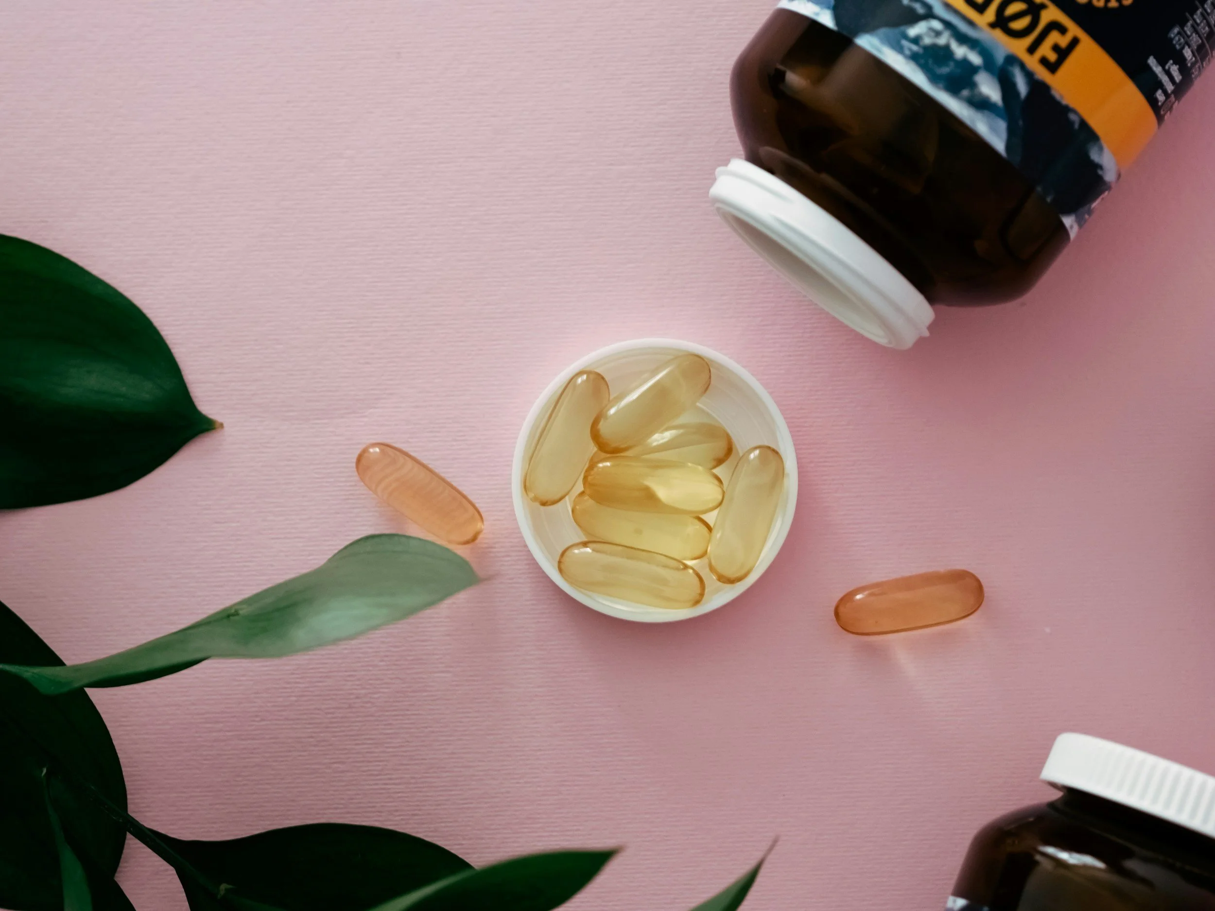 A pink background with green leaves in the lower left corner, a small white bowl filled with yellowish gel capsules, and two brown bottles with white caps and partially visible labels on the top right.