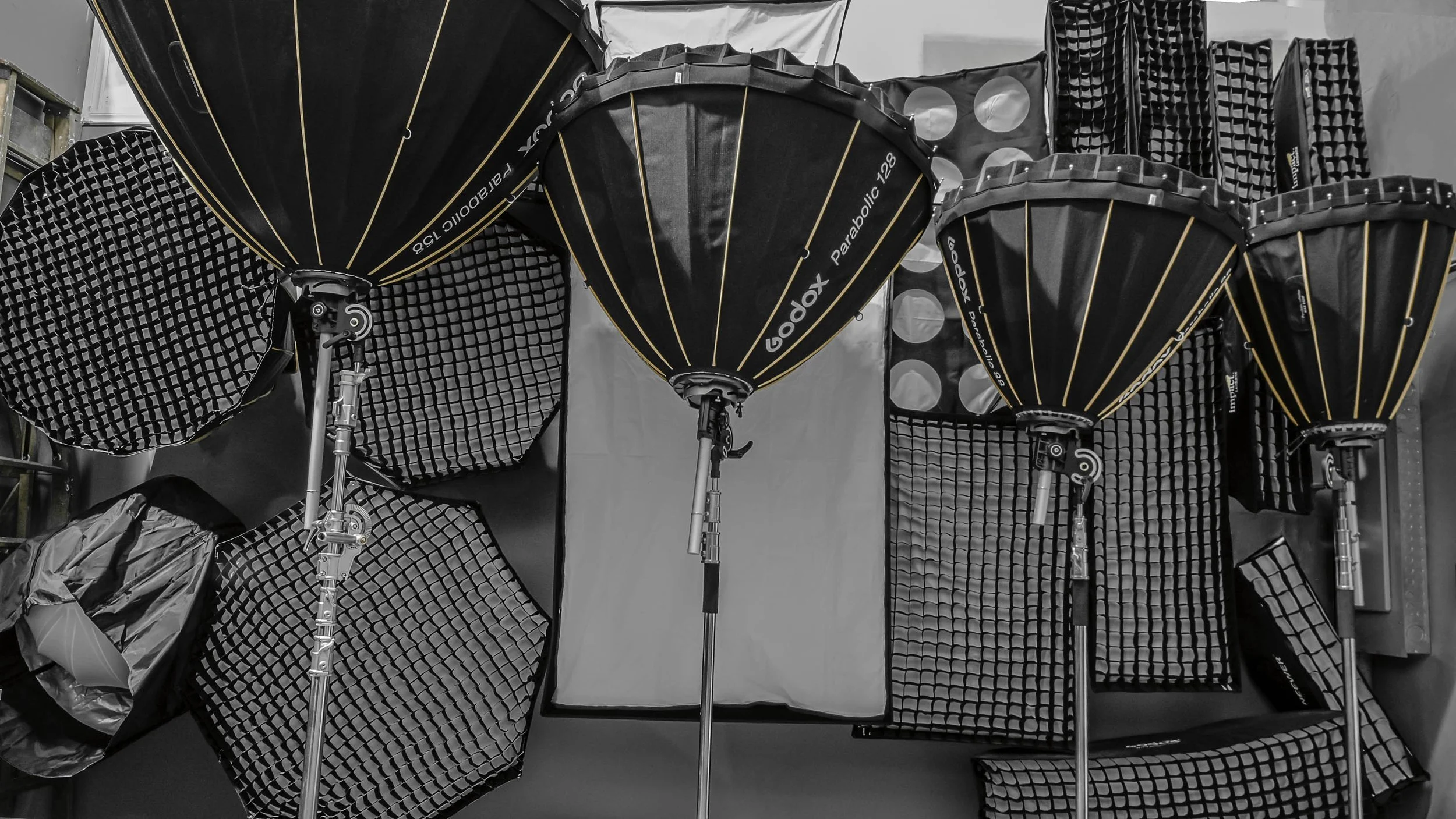 A collection of professional studio lighting equipment, including four large black umbrellas with orange accents, some grid diffusers, and a white reflector, arranged on a table or floor.