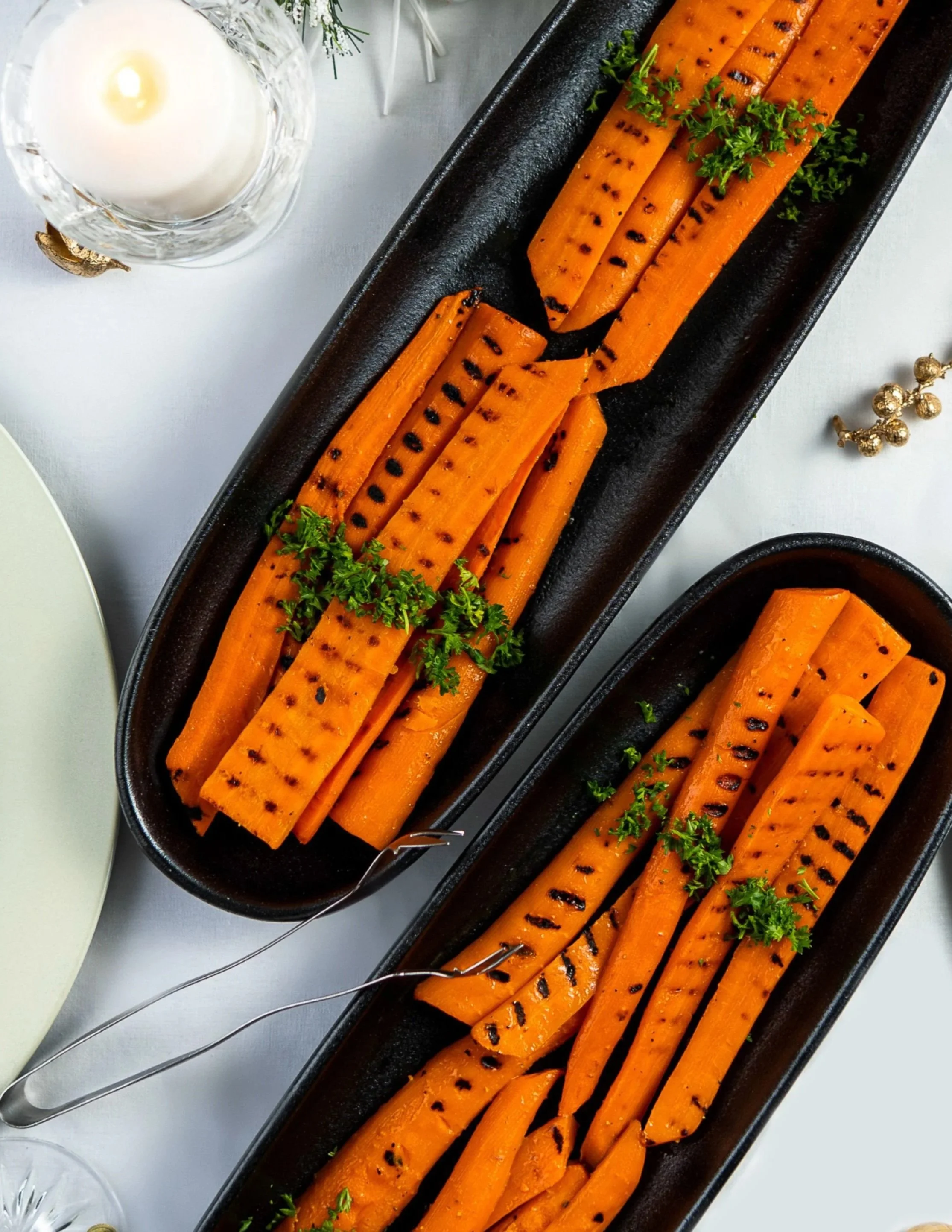 Grilled carrots garnished with parsley on a black rectangular plate, with a glass of white wine and some holiday decorations nearby.