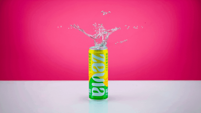Can with colorful branding, open at the top with water splashing out, placed on a white surface against a pink background.
