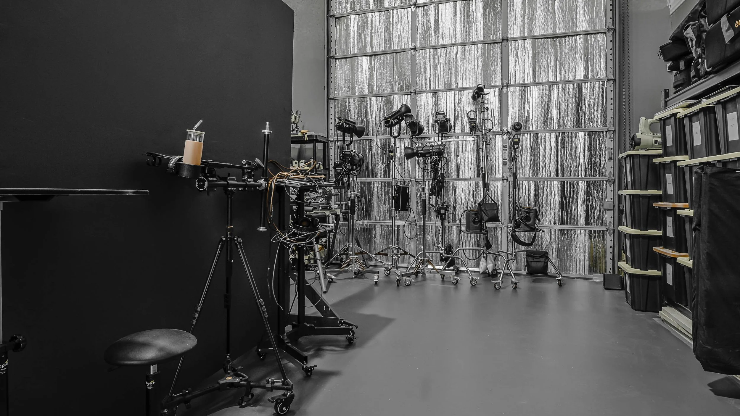 A photography studio with professional lighting equipment and a black backdrop, including light stands, softboxes, and other gear organized on rolling carts against a metallic background.