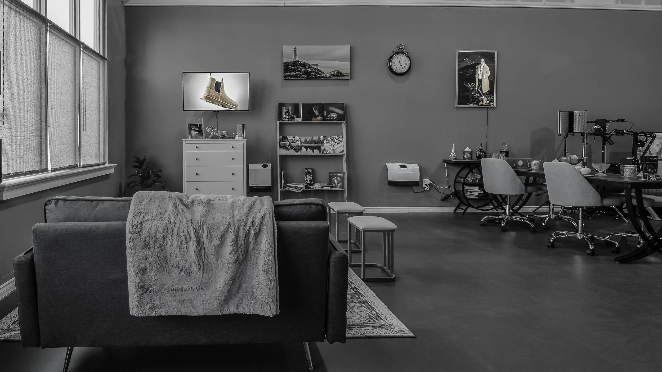 A gray and white living room with a couch, a side table, a white dresser, and a picture of a lighthouse on the wall. There are several framed pictures, a clock, and a TV on the wall. The room also has a dining table with chairs, and shelves with vari