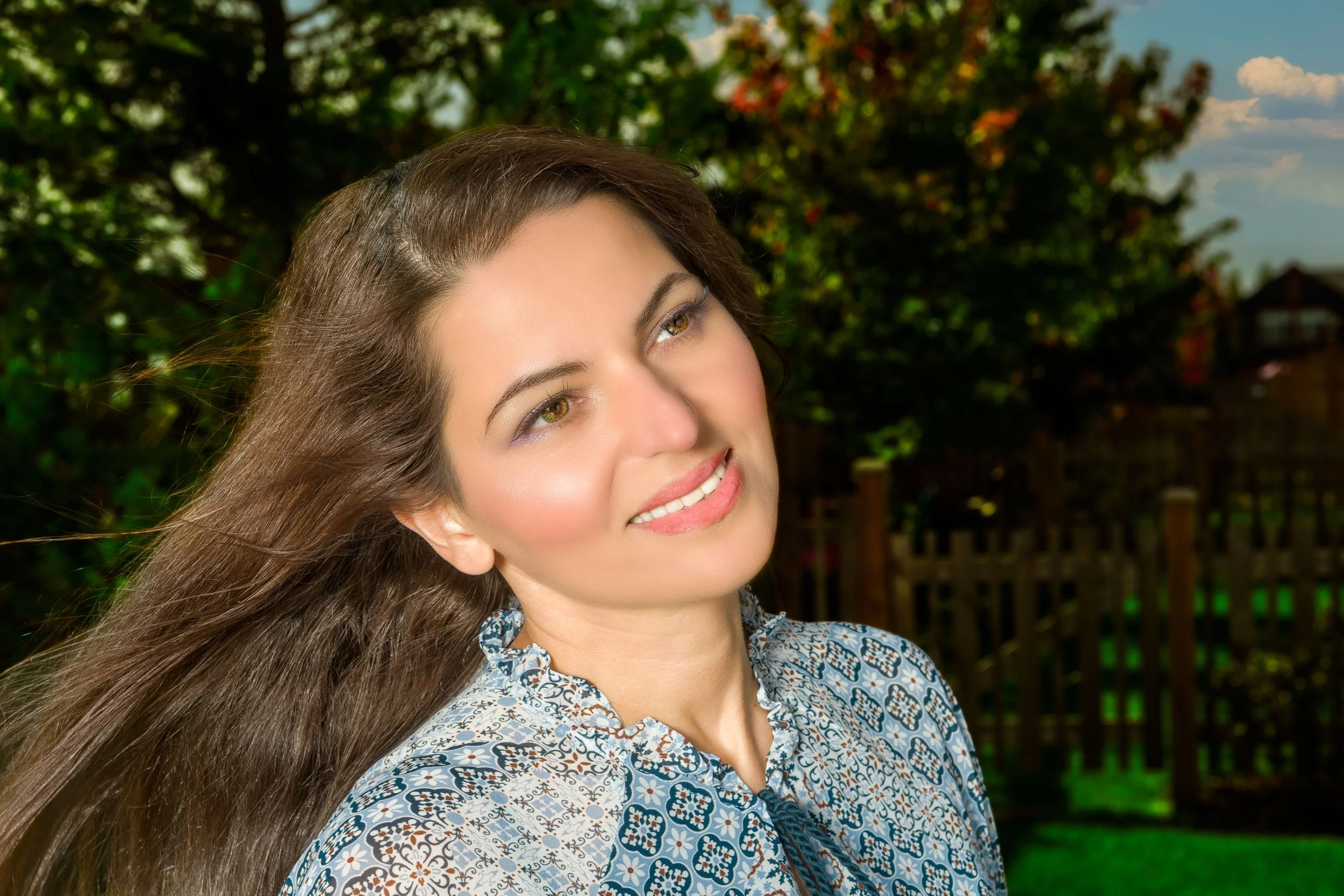 A woman with long brown hair smiling outdoors with trees and a wooden fence in the background.