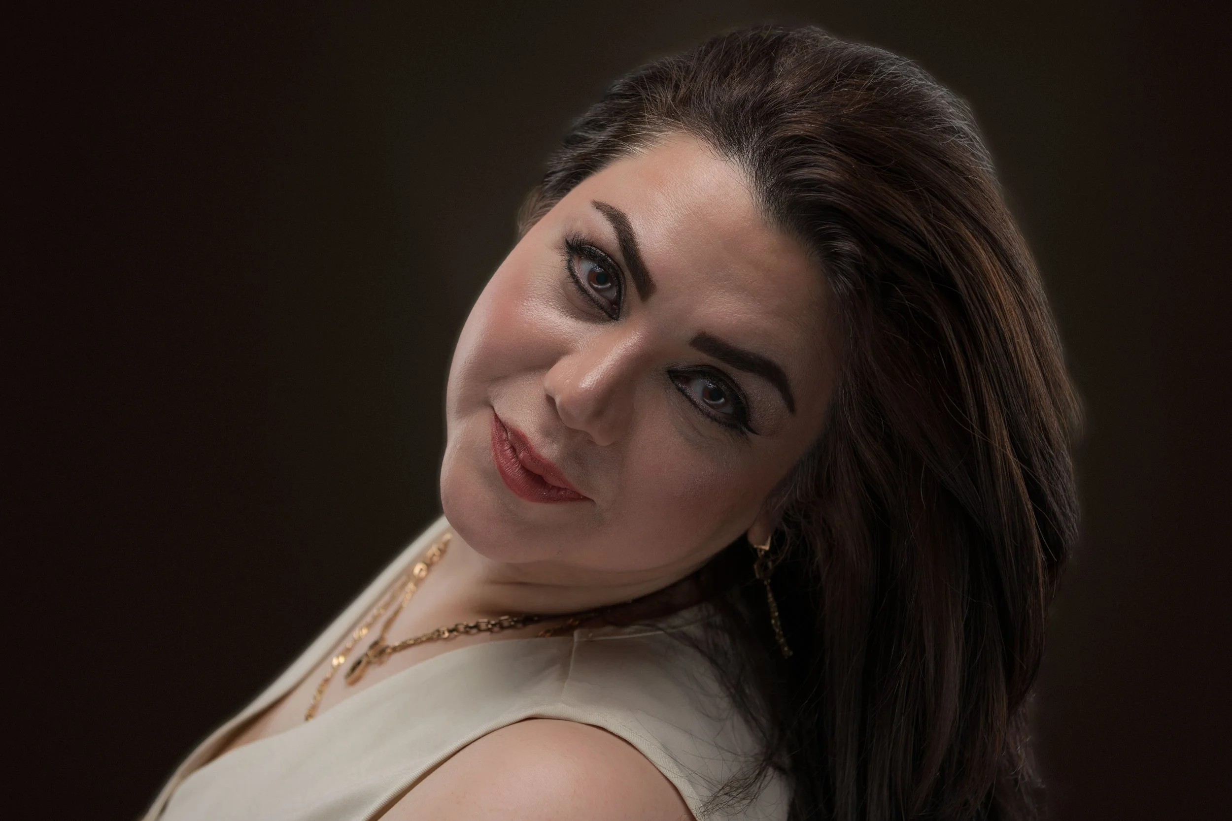 A woman with dark brown hair, blue eyes, and makeup, wearing a cream-colored dress and gold jewelry, smiling softly at the camera against a dark background.