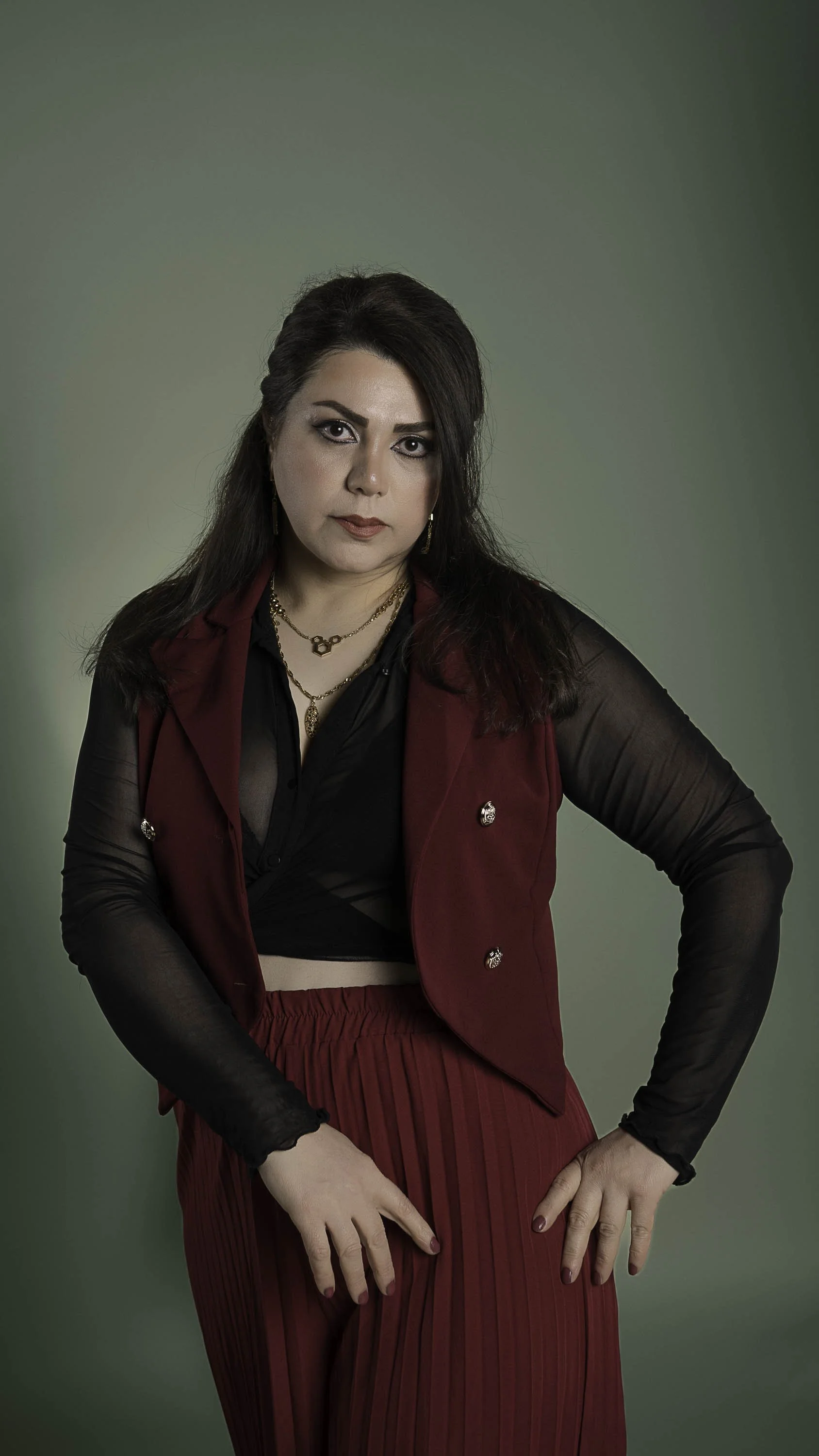A woman with dark hair, wearing a black sheer top layered with a red vest and a matching pleated red skirt, standing against a green background.