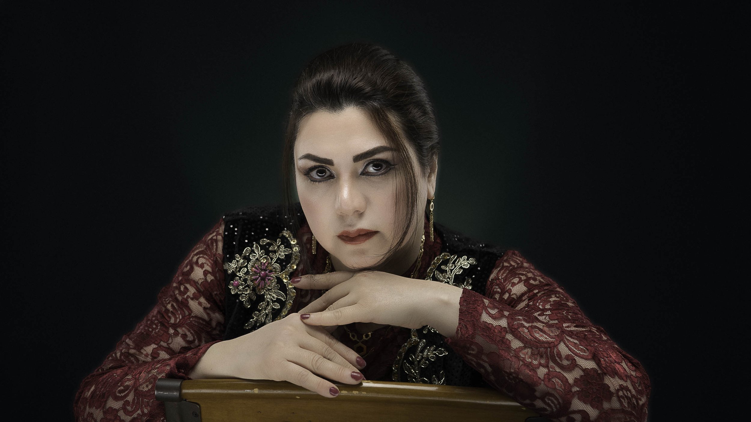 A woman with dark hair styled back, wearing a red lace dress and a black velvet vest with gold embroidery, poses with her hands resting on a wooden surface against a dark background.