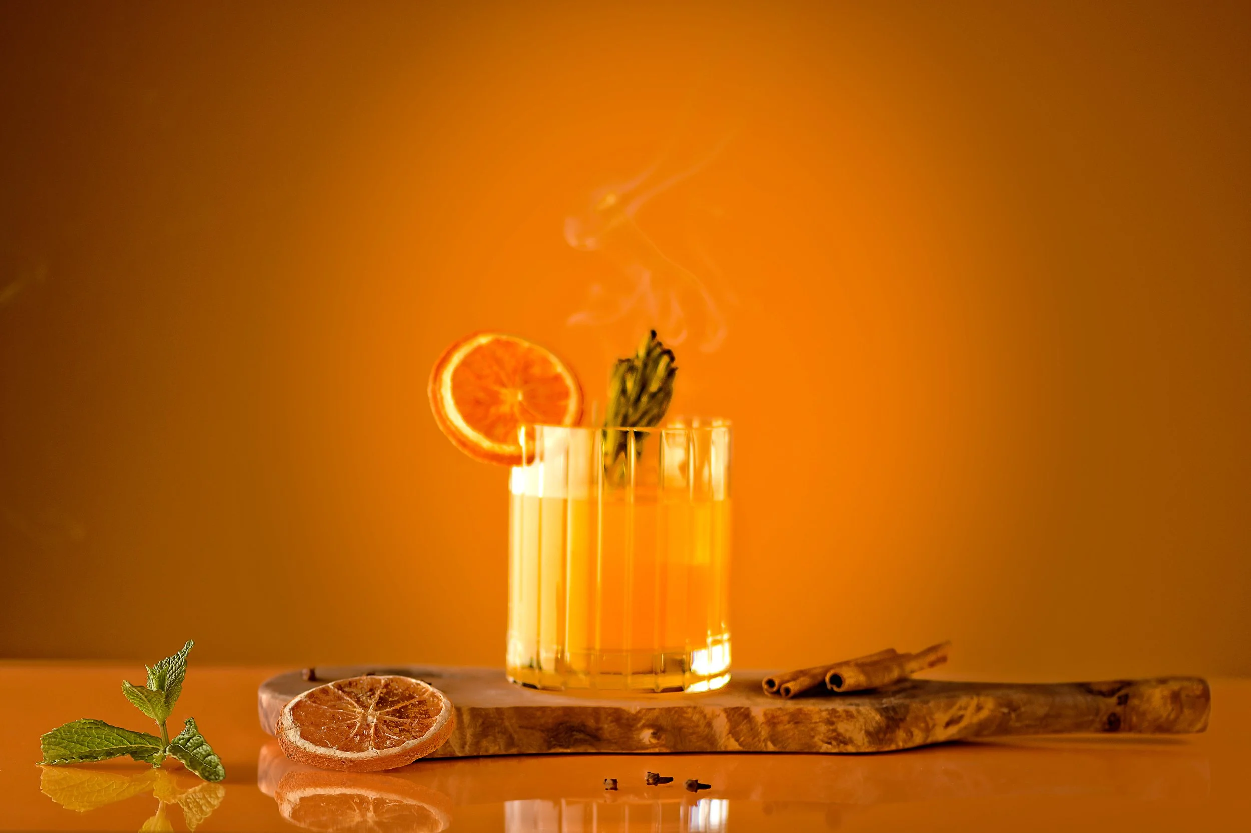 A glass of orange-colored beverage garnished with a slice of orange and a sprig of herbs, placed on a wooden surface with dried citrus slices, mint, cinnamon sticks, and peppercorns, with a warm orange background.