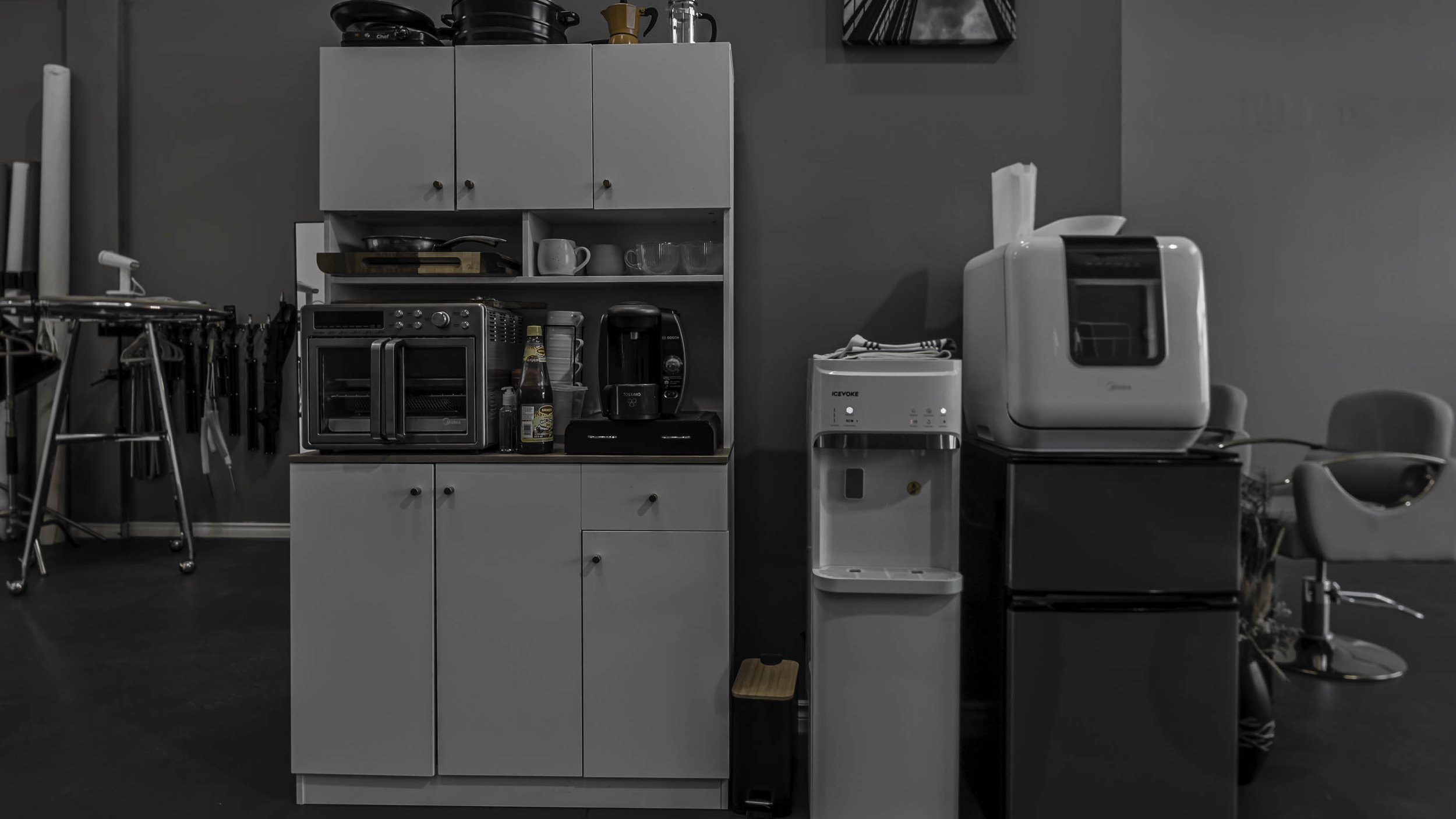 Kitchen area with various appliances including a toaster oven, coffee maker, and water dispenser, alongside kitchen chairs and cabinets.