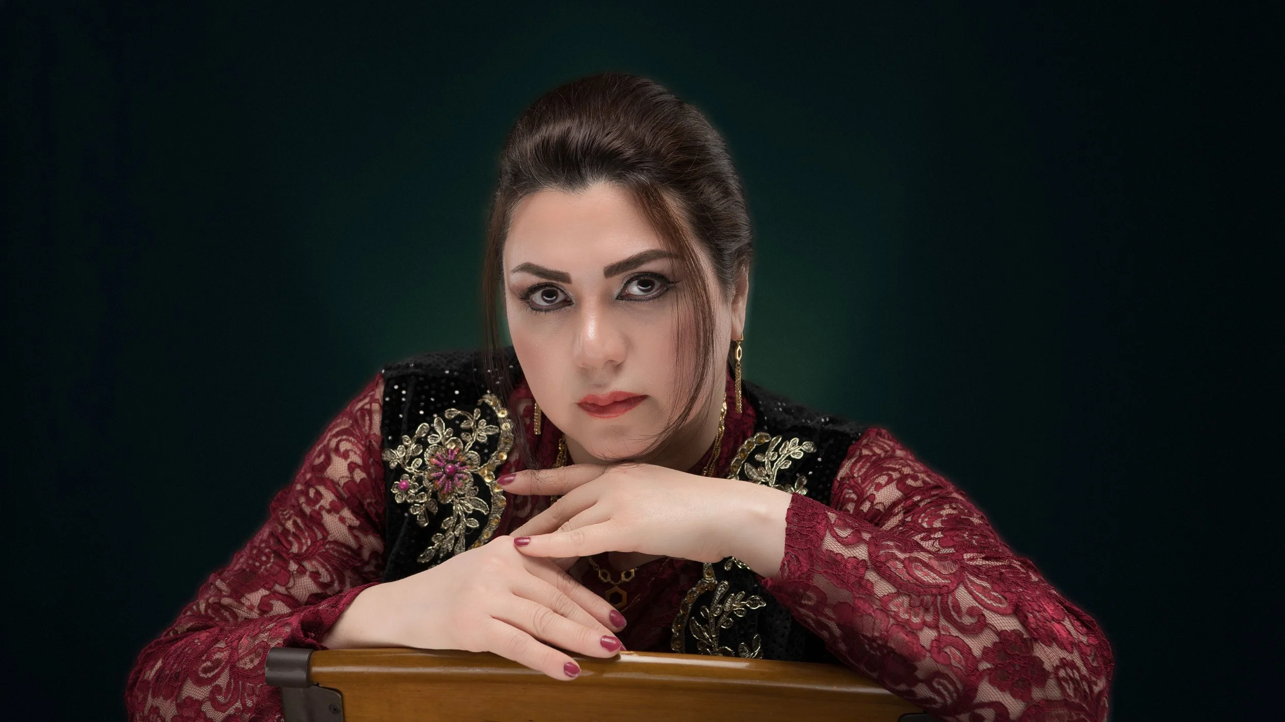 A woman with dark hair styled back, wearing a red lace dress and a black velvet vest with gold embroidery, poses with her hands resting on a wooden surface against a dark background.