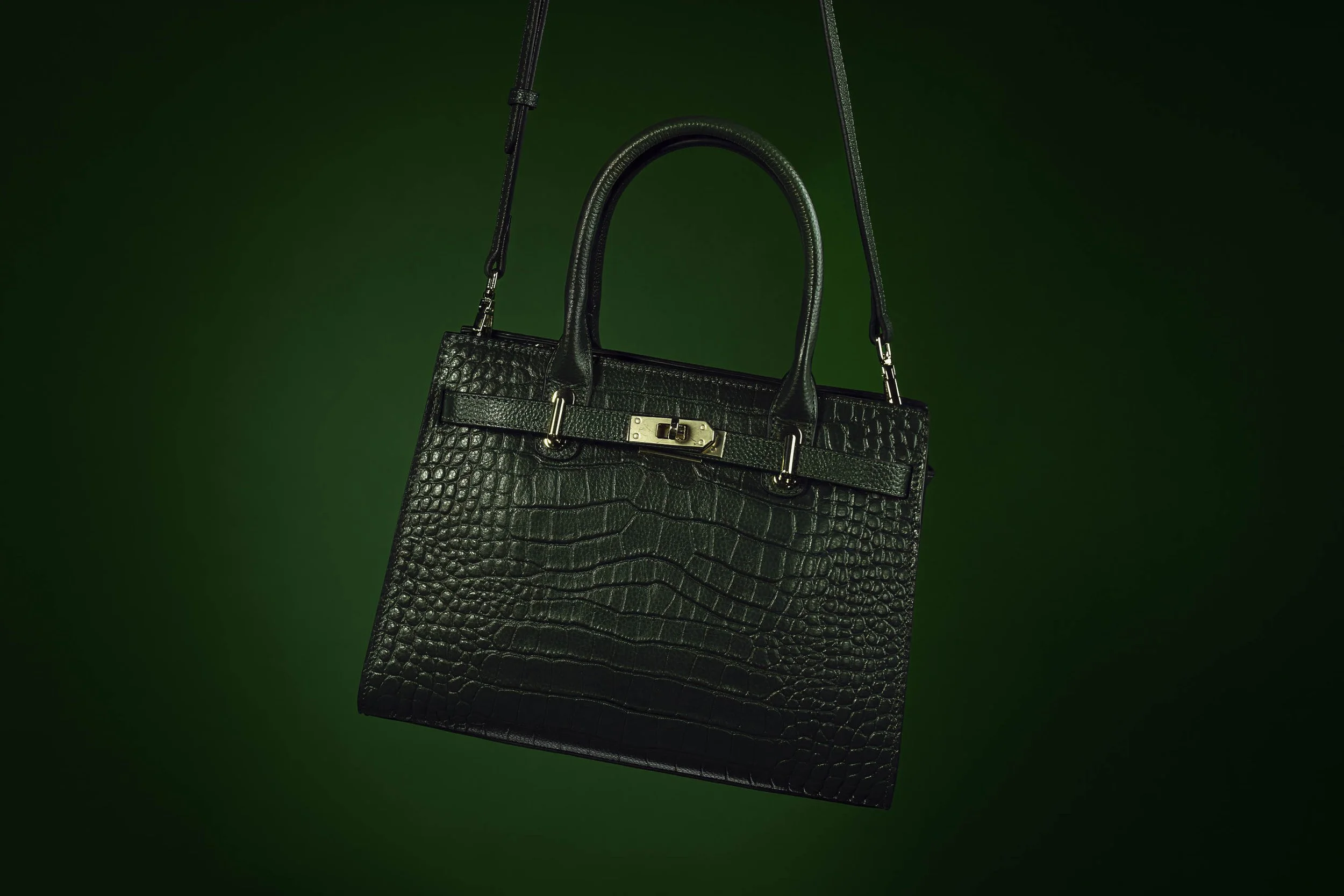 A black crocodile leather handbag with gold hardware and a detachable shoulder strap, hanging against a green background.