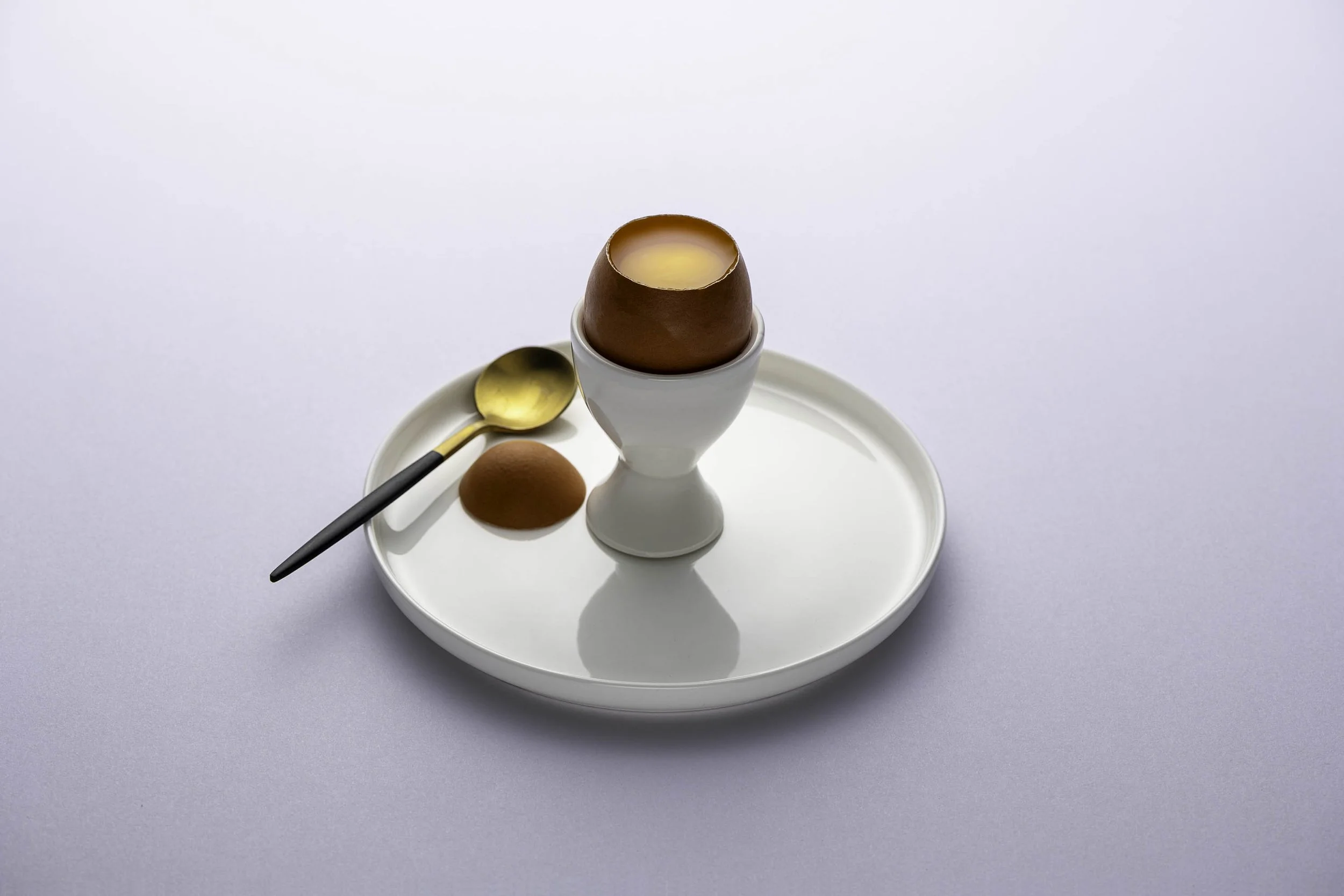 Egg in a cracked eggshell inside an egg cup, with a spoon and a whole egg on a white tray.