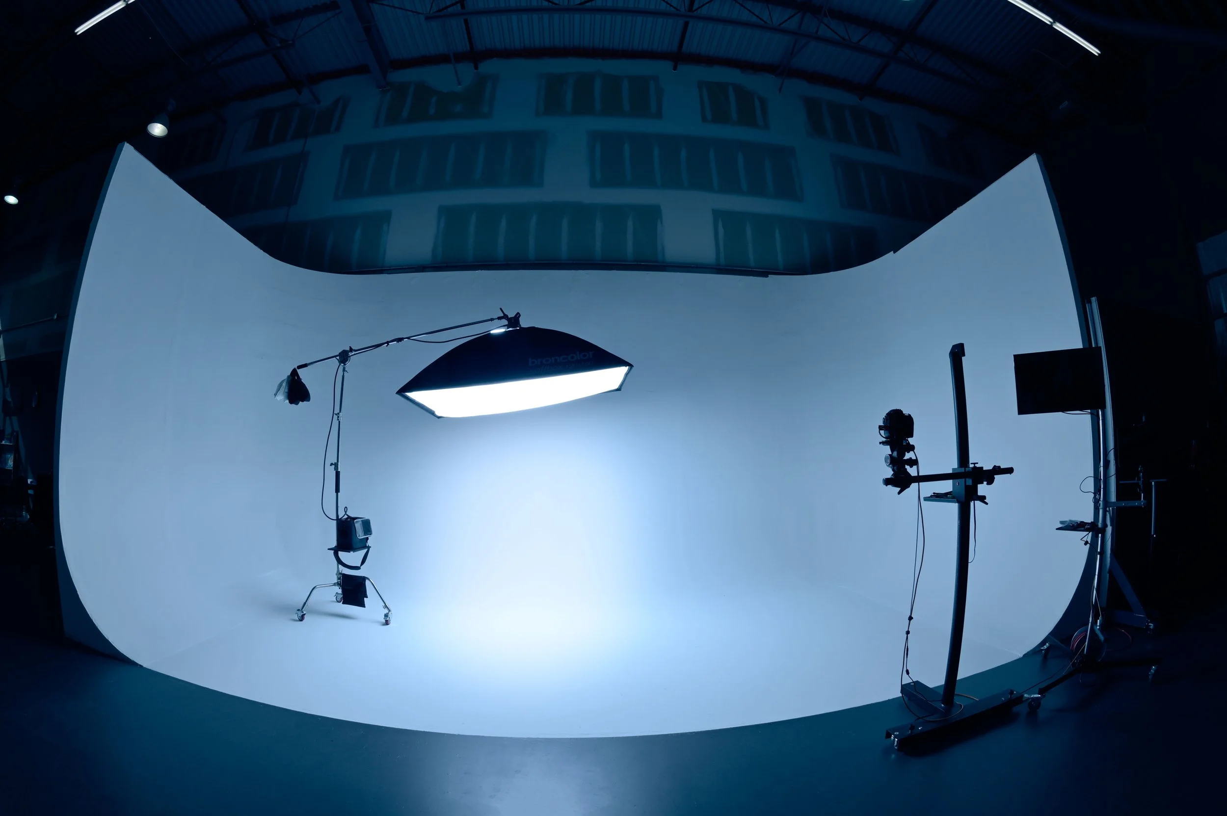 Photography studio with a white cyclorama wall, professional lighting equipment including two softbox lights, a camera on a tripod, and other studio gear, set up for photo or video production.