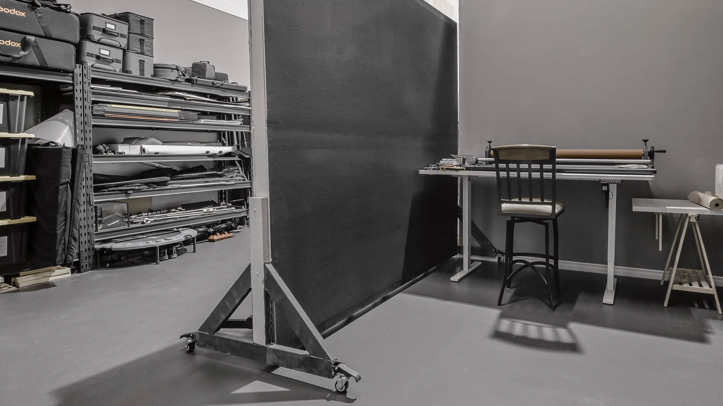 A photography studio or art workspace featuring a large blackboard on wheels, a black chair, a drafting table with a roll of brown paper, and shelves of photography equipment and props in the background.