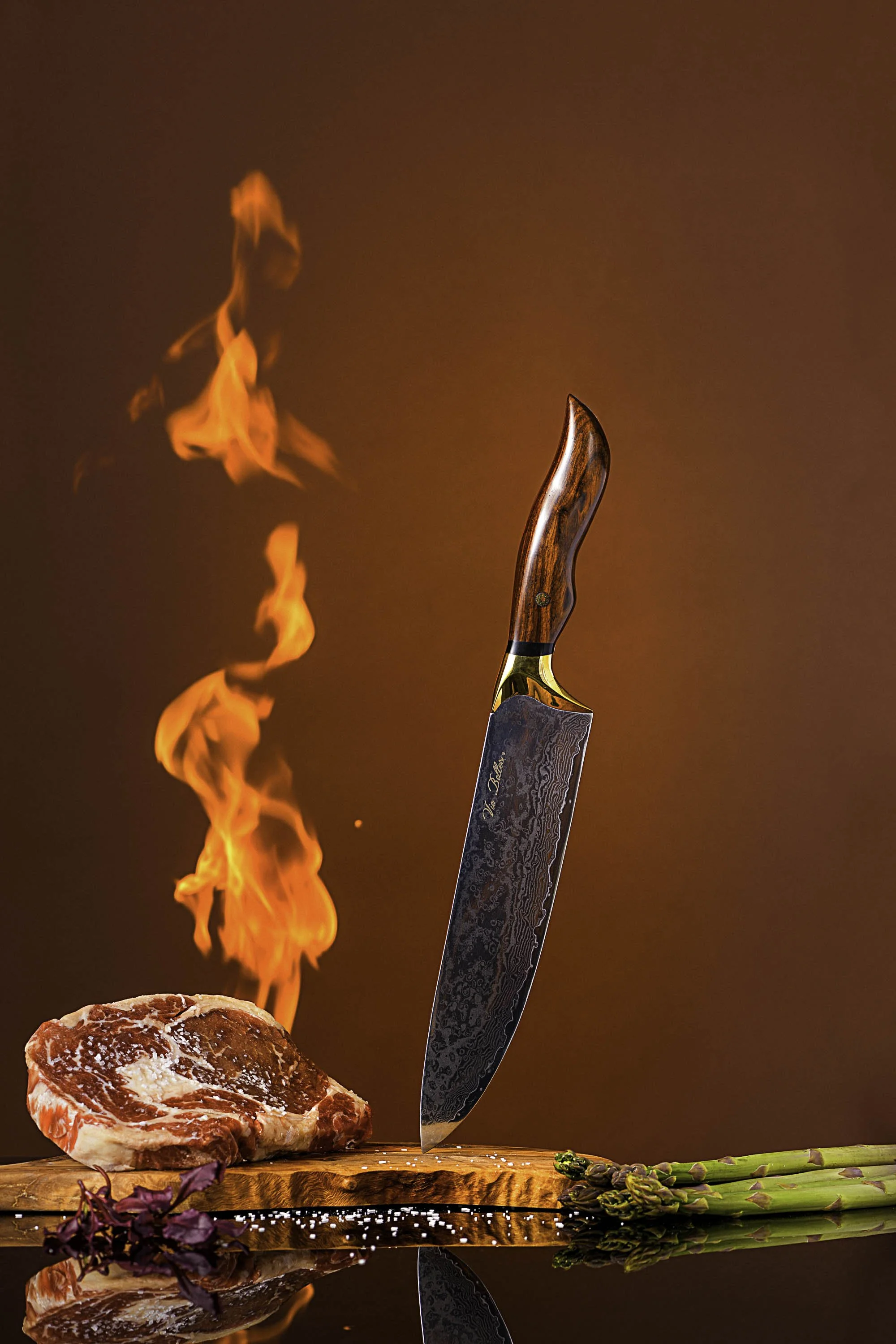 A high-quality image showing a chef's knife, with a Damascus pattern, stabbing a raw steak on a wooden cutting board. Fire is rising from the steak, creating a dramatic effect. There are fresh asparagus spears lying beside the steak. The background i