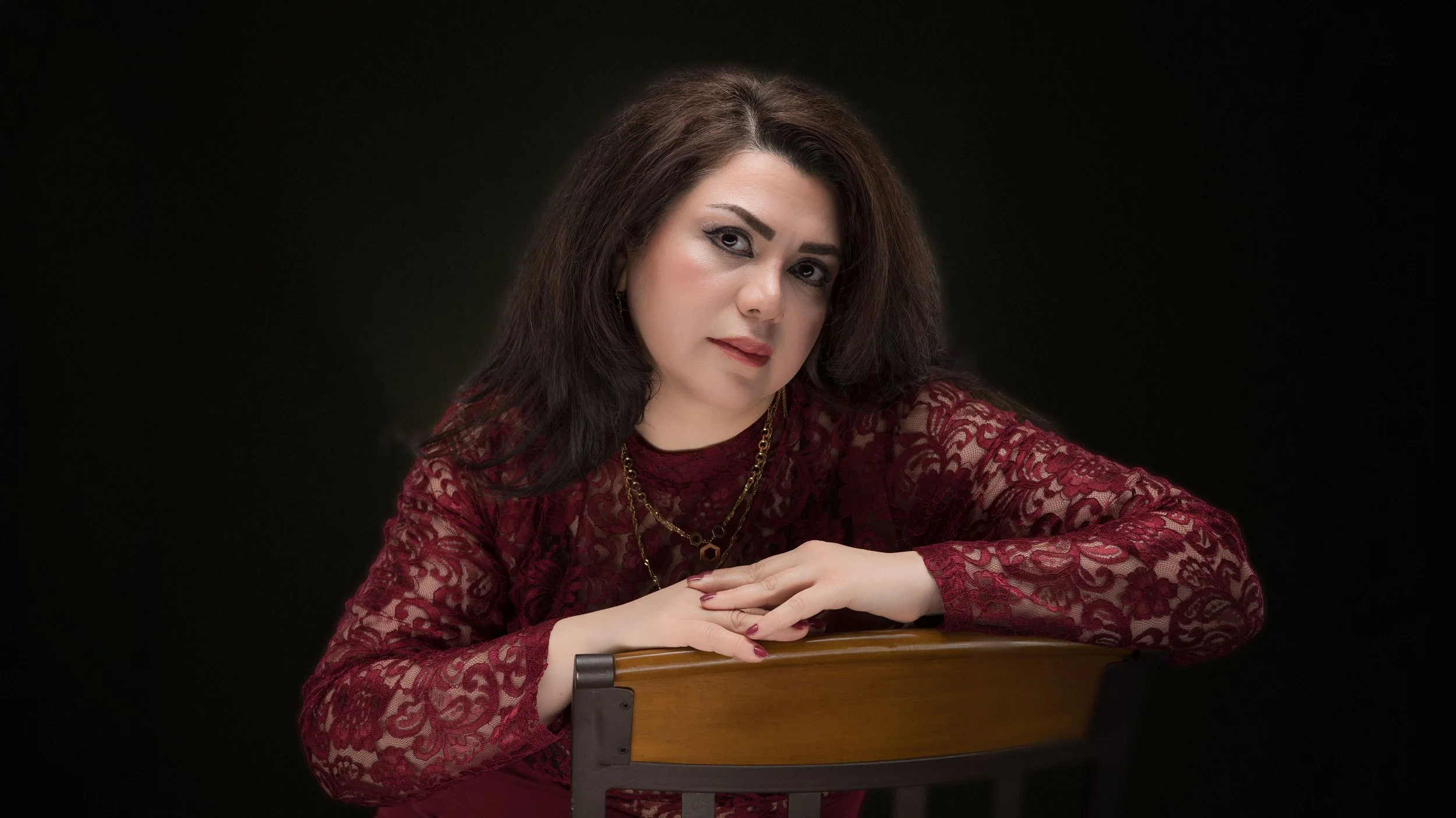 Woman with dark brown hair in a burgundy lace top, gold necklaces, and makeup, leaning on a wooden chair against a black background.