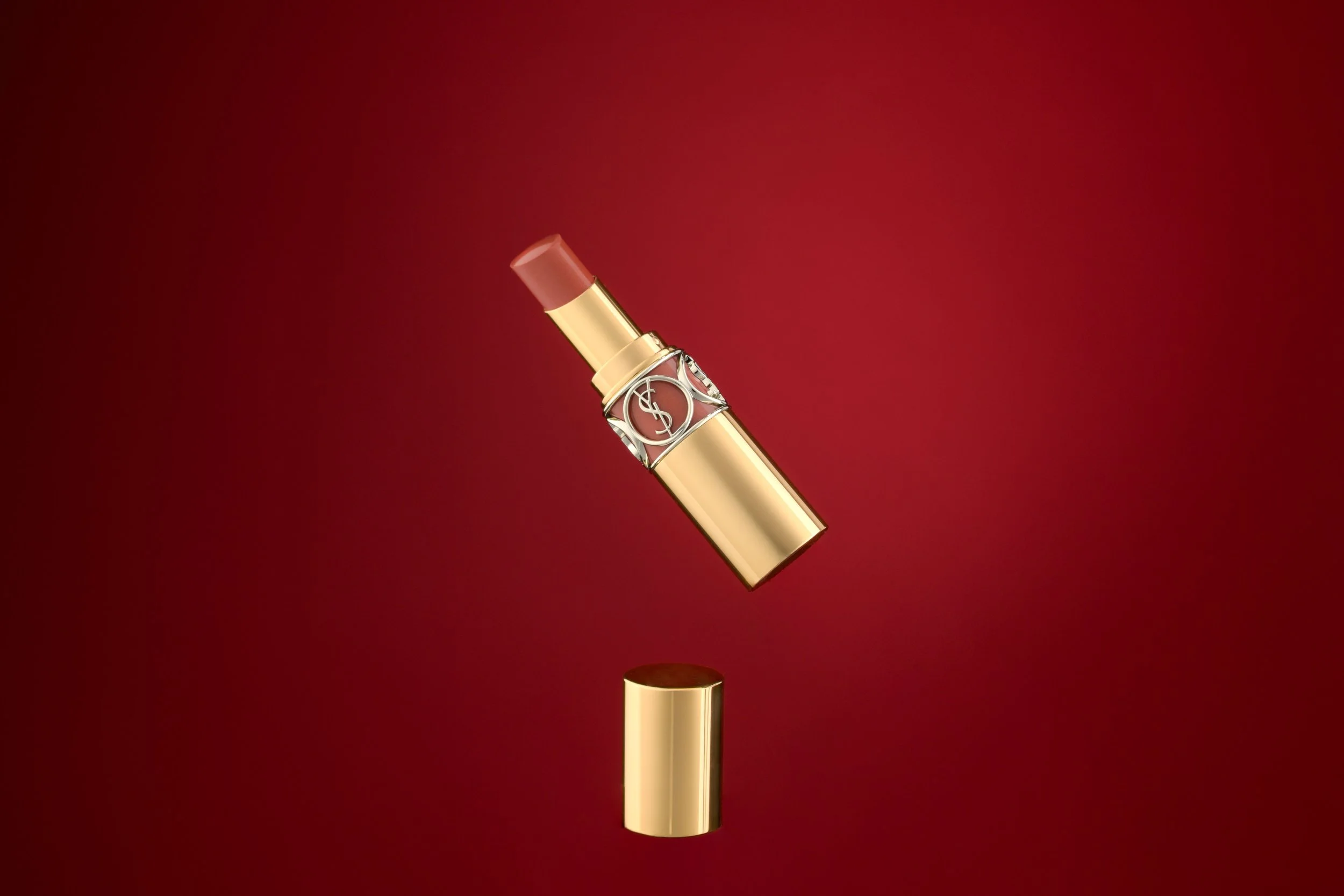 A gold lipstick with a pinkish-brown shade is breaking in half, with the top part floating above the bottom part against a red background.