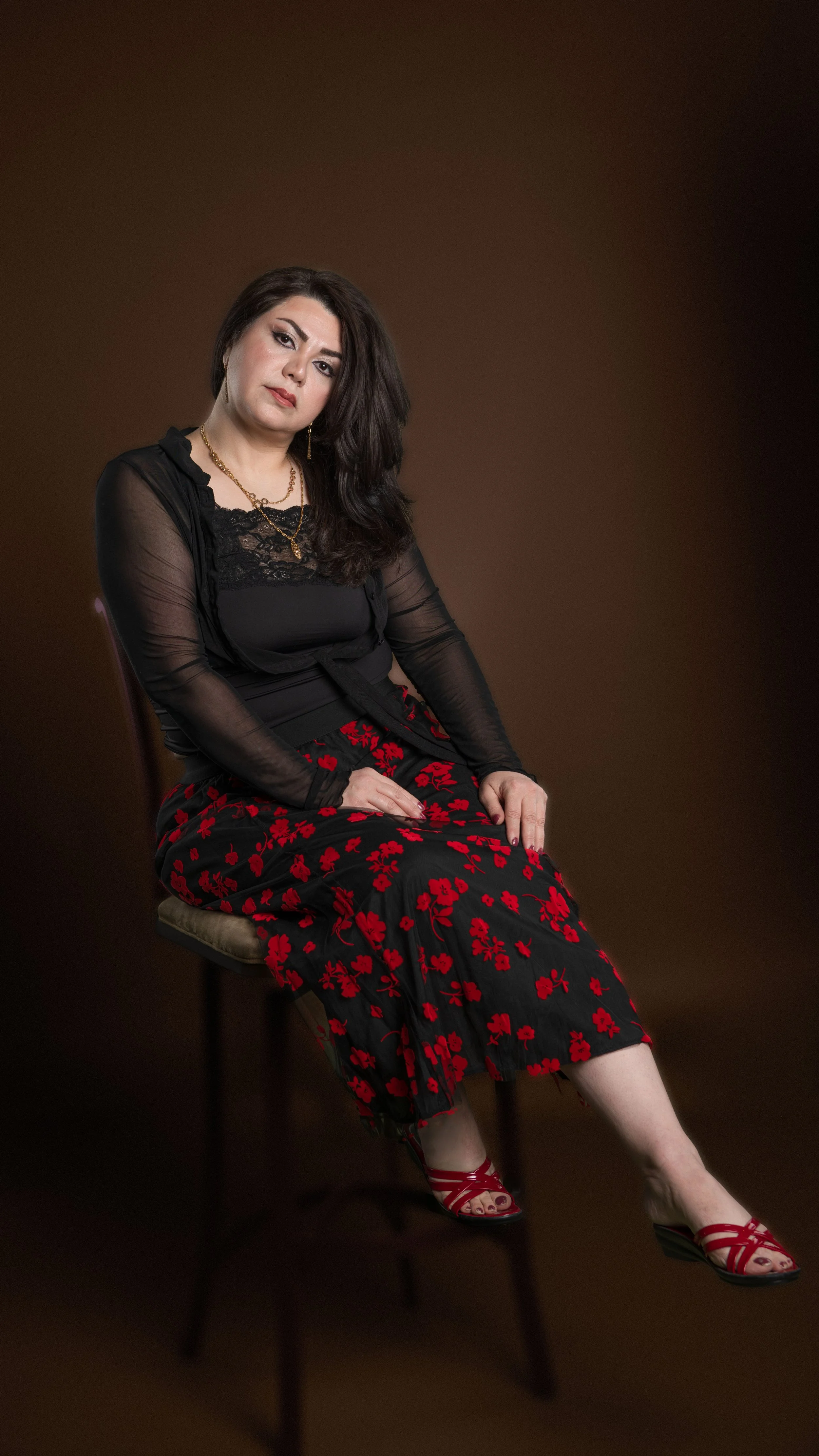 A woman with dark hair and makeup, wearing a black lace top, sheer black jacket, red and black floral skirt, and red sandals, sitting on a chair against a brown background.