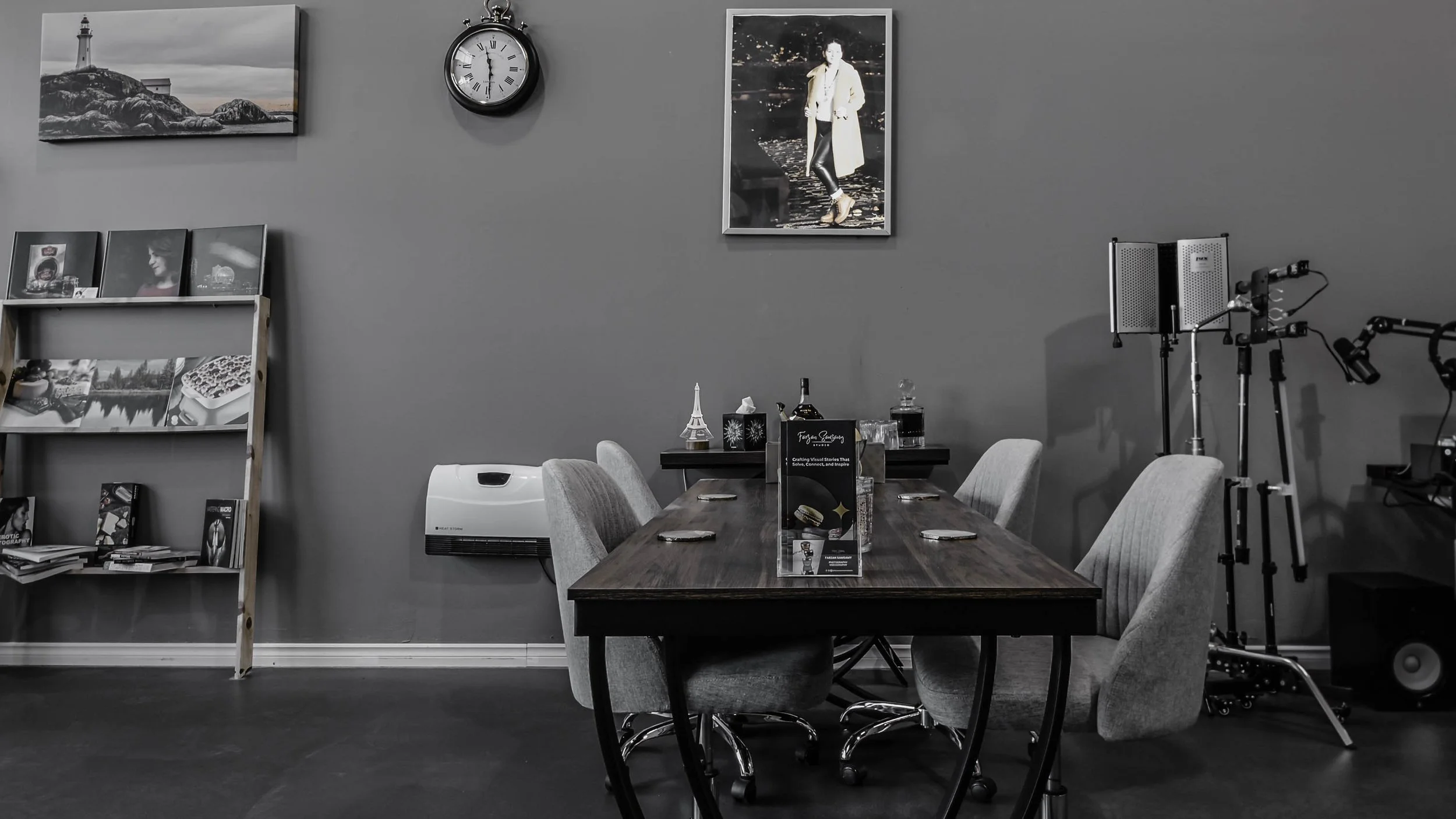 A conference room with a wooden table surrounded by six gray upholstered chairs, a gray wall decorated with black and white framed photographs and a large portrait, a clock showing 10:09, a white air purifier on the wall, a shelf with black and white