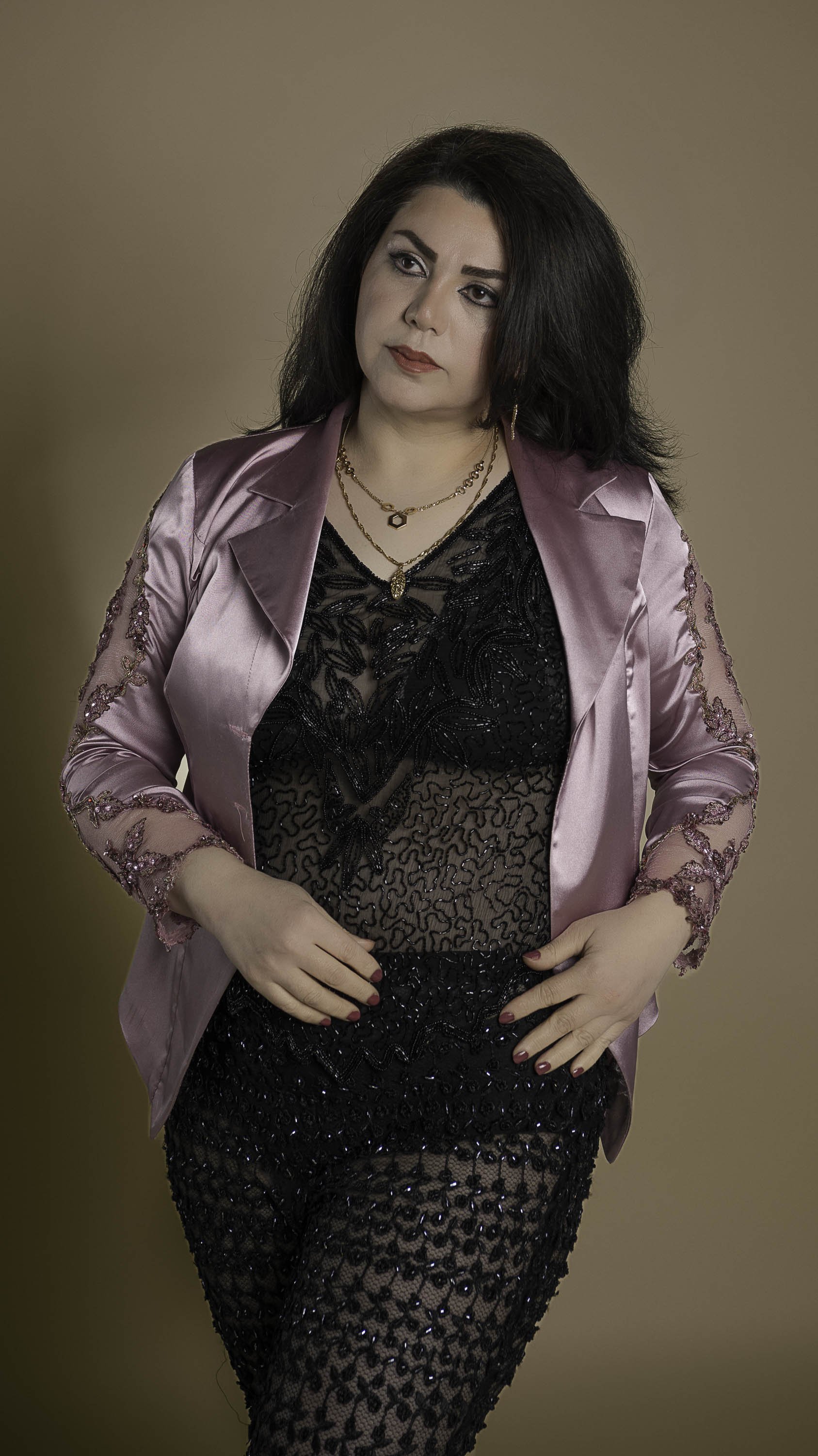 A woman with dark hair and makeup wearing a pink satin blazer over a black lace top and leopard print sequined skirt, standing against a beige background.