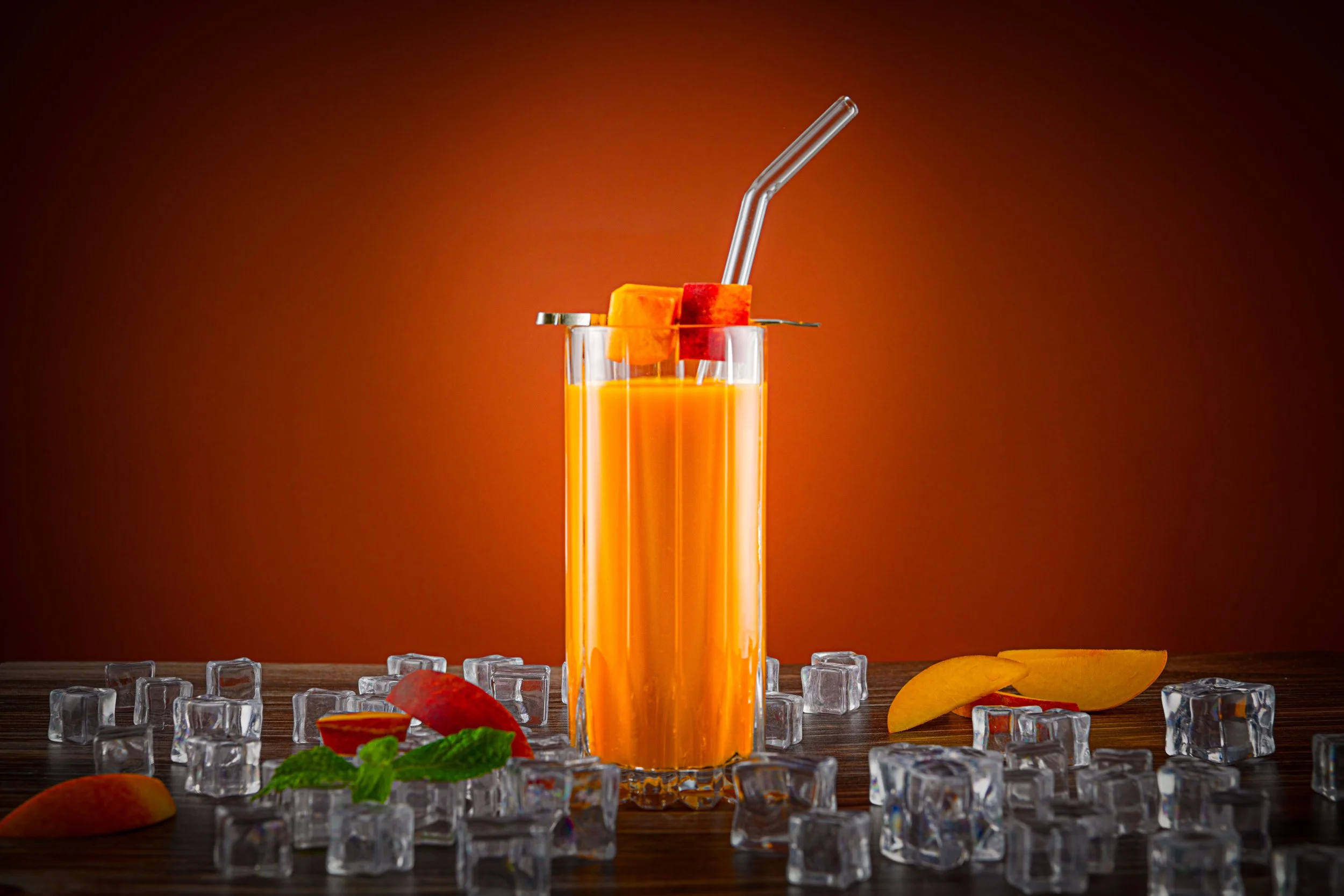 A tall glass of orange juice with ice cubes, garnished with peach and watermelon pieces, on a wooden surface with scattered ice cubes, slices of peach, and mint leaves, against a dark gradient background.