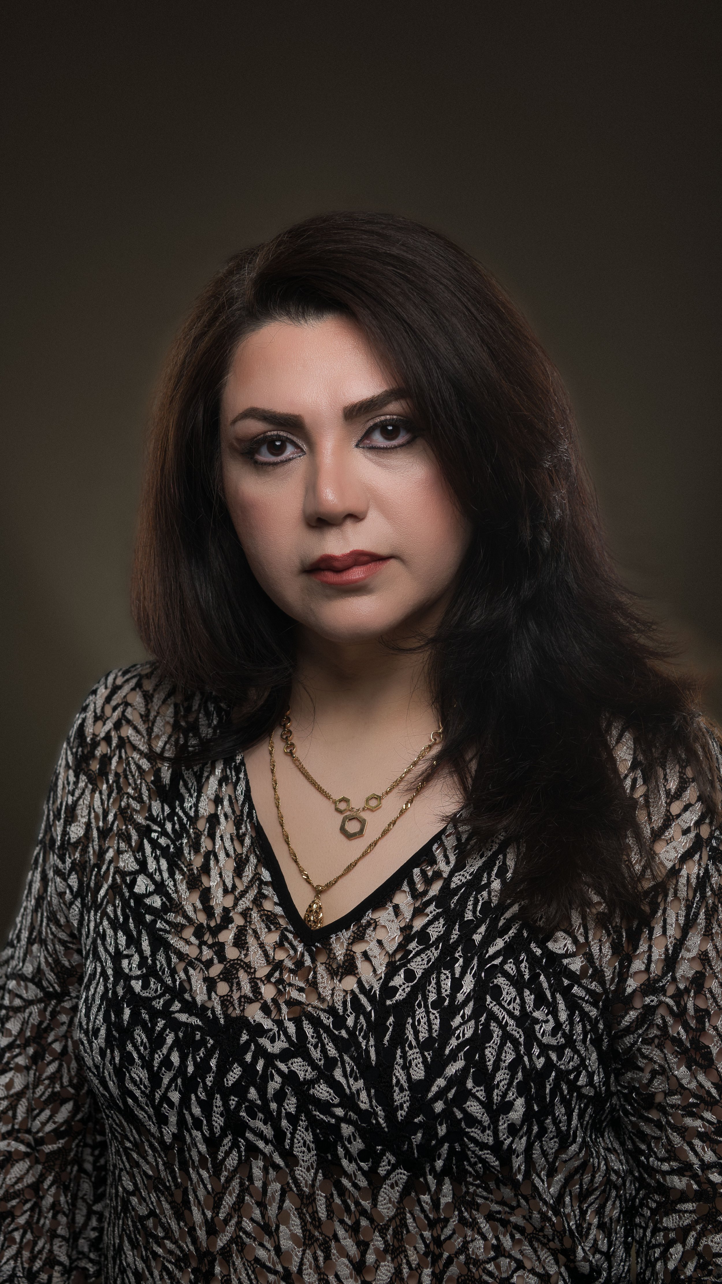 Portrait of a woman with shoulder-length dark hair, wearing a patterned blouse and layered gold necklaces, looking at the camera against a dark background.