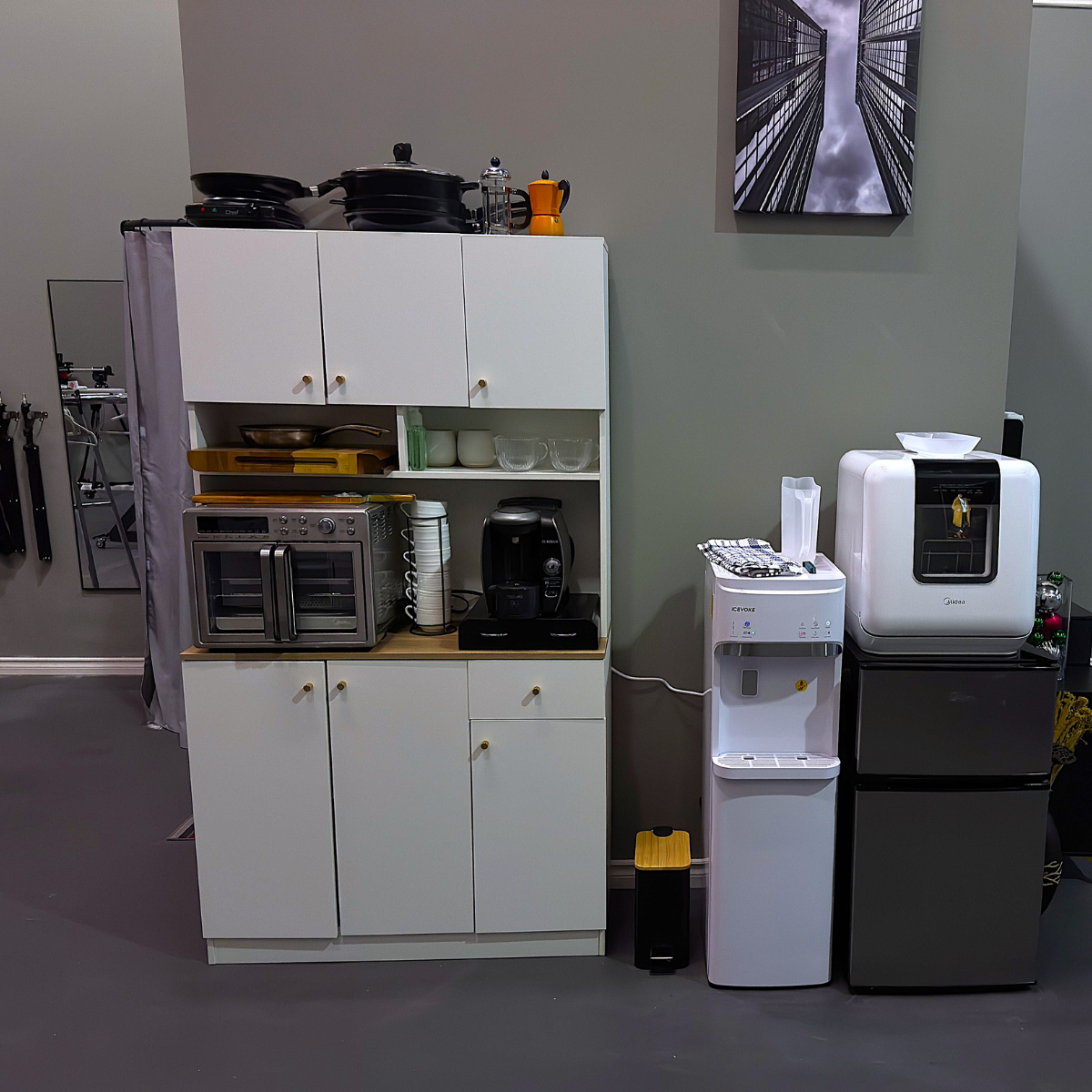 A kitchenette area with a white cabinet, a microwave, a coffee machine, and a water dispenser. To the right, a water cooler and a mini refrigerator with a white appliance on top, possibly a microwave or a small oven. A wall-mounted picture of tall bu