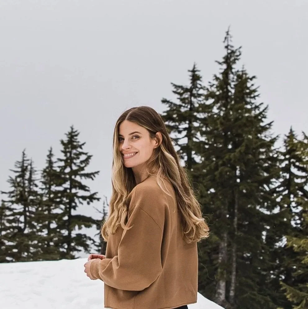 A woman with long blonde hair smiling outdoors in a snowy landscape with evergreen trees.