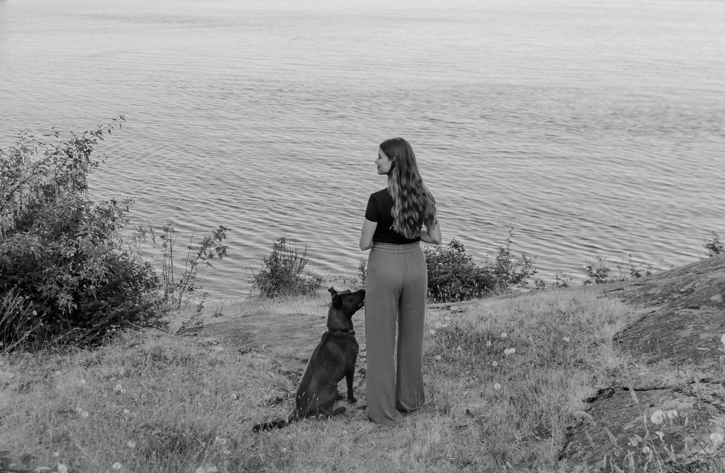 A woman with long wavy hair in a dark top and wide-leg pants stands on grass near a body of water, looking away, with a dog sitting beside her.