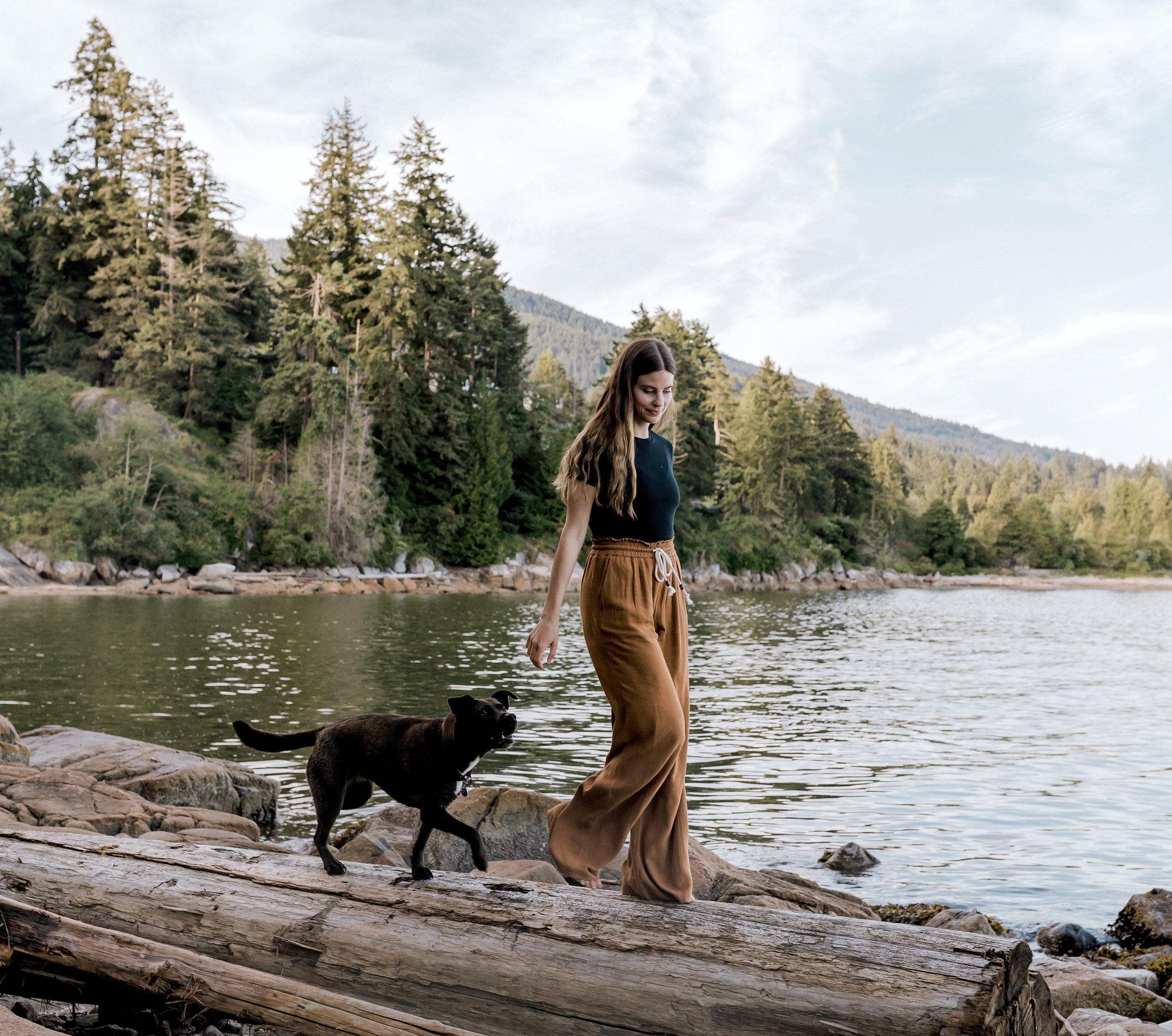 A young woman with long brown hair walking on a fallen log at the edge of a lake, with a black dog nearby, surrounded by pine trees and mountains in the background.