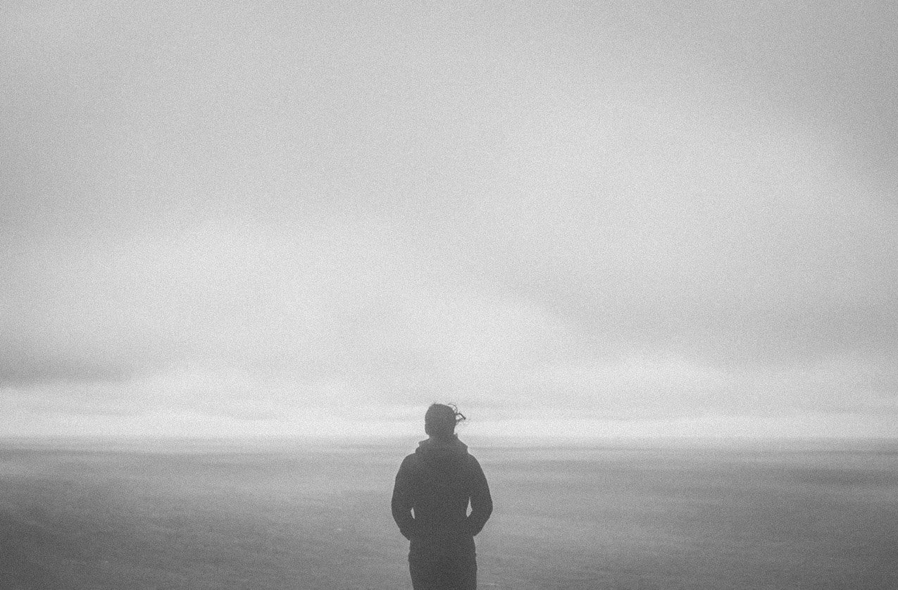 Person standing alone on a beach, facing the ocean, in black and white.