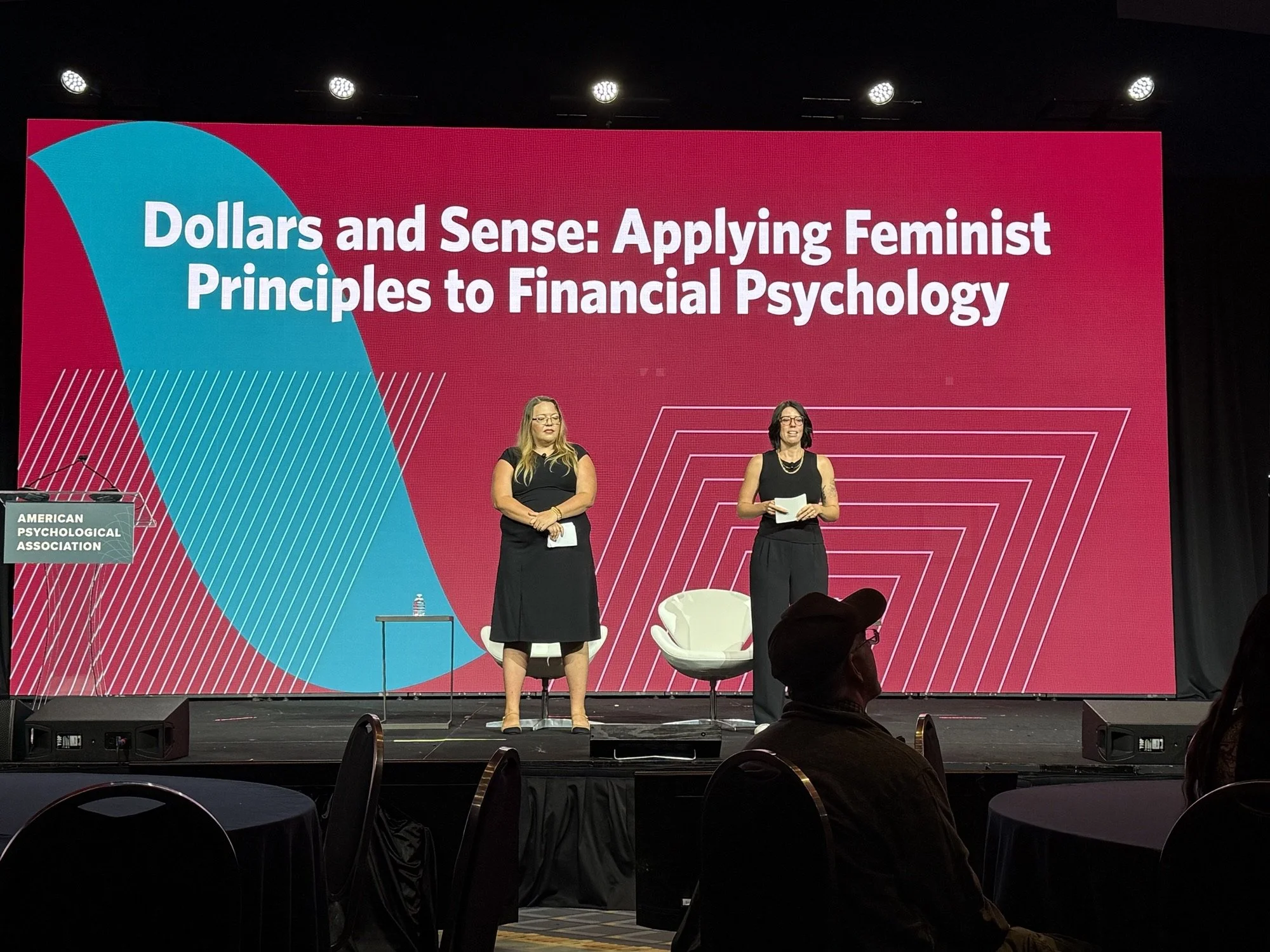 Two women standing on a stage in front of a large red and blue sign that reads 'Dollars and Sense: Applying Feminist Principles to Financial Psychology.' One woman holds papers, the other stands with her hands clasped in front. Audience members are visible seated at tables in front of the stage.